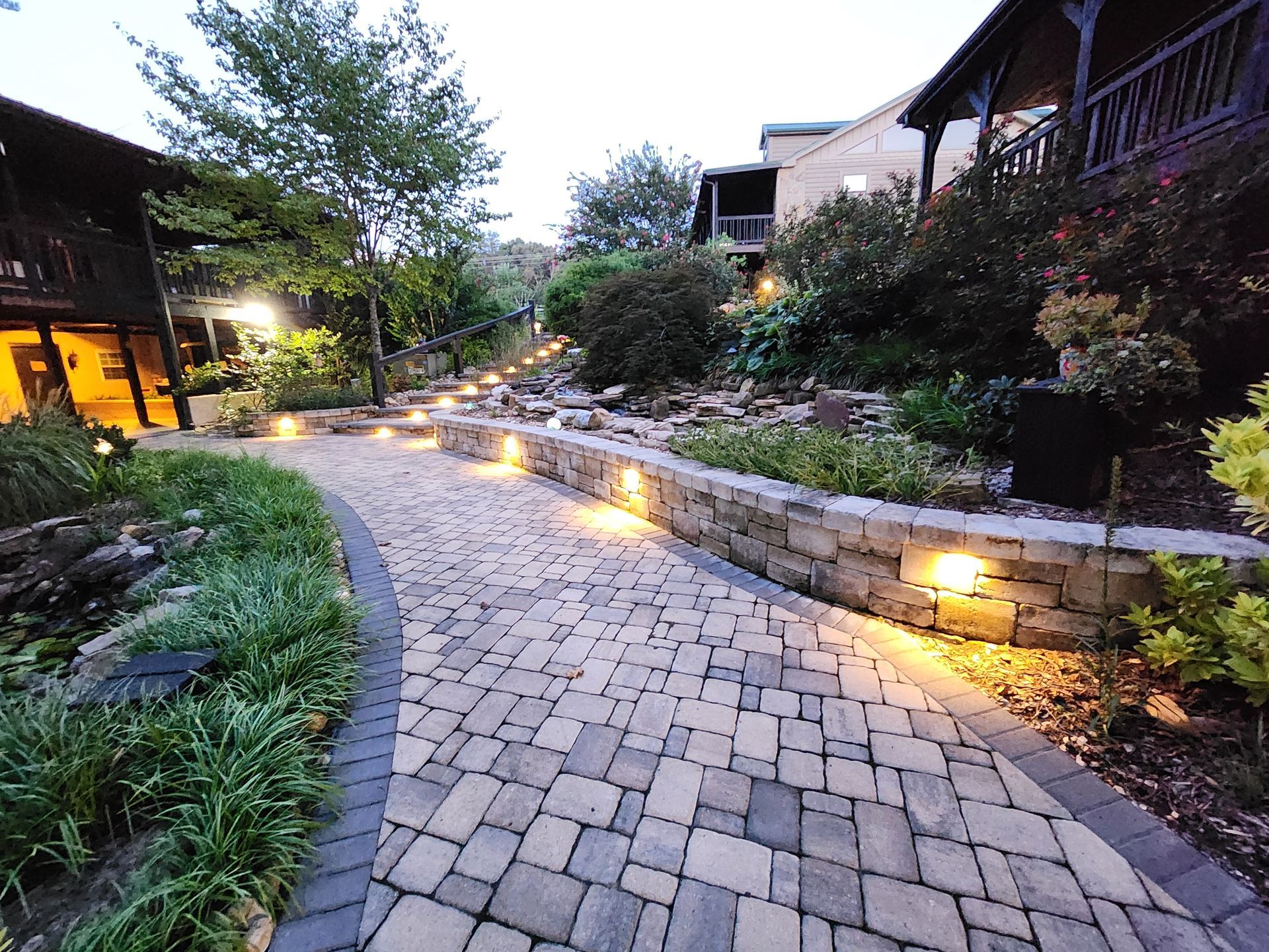 Brick pathway lined with lights, leading through landscaped yard. Buildings in background.
