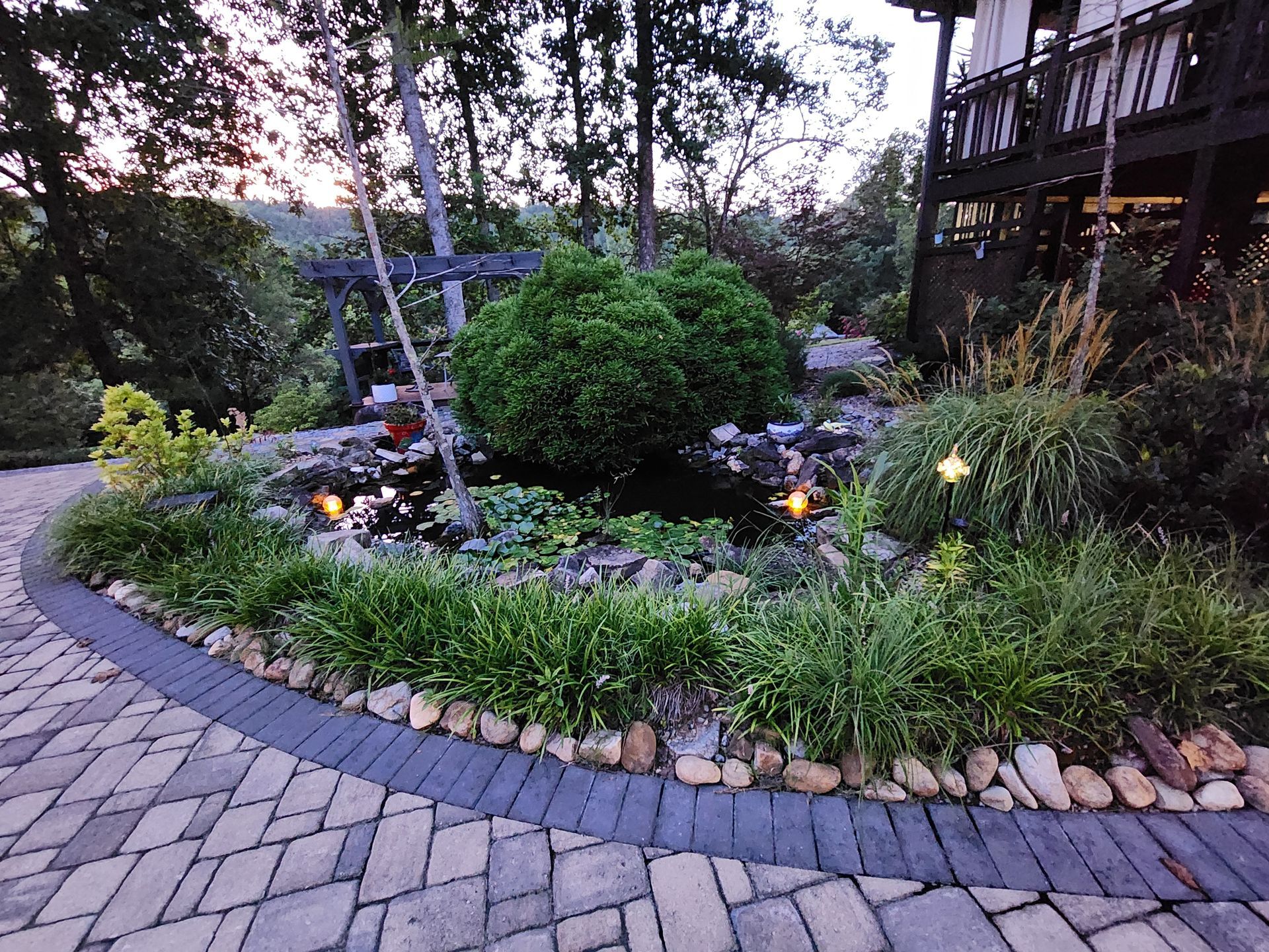 Stone patio surrounds a lush garden bed with a small pond, shrubs, and ornamental plants.