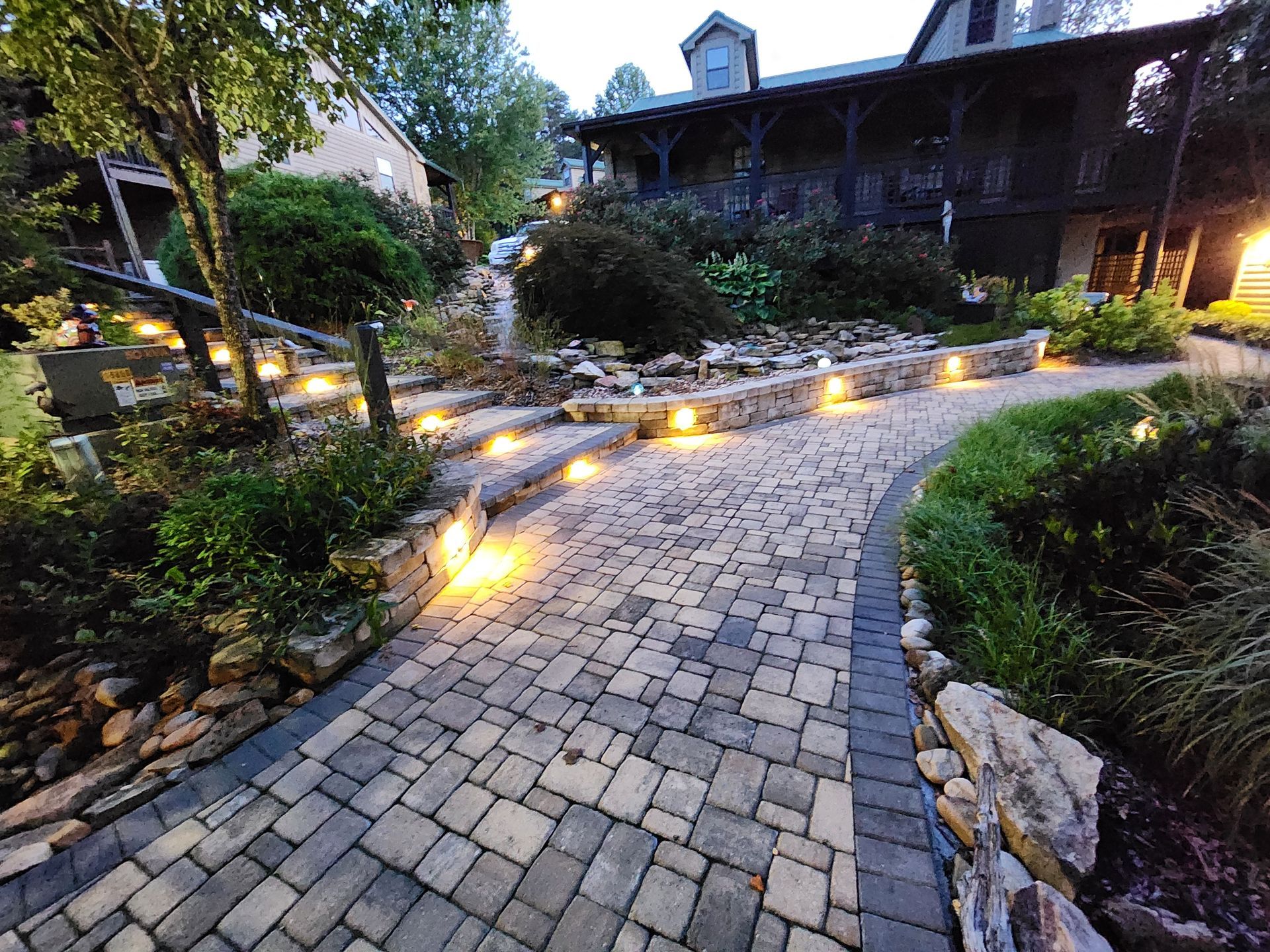 A brick pathway lit by small lights leads to a multi-level building surrounded by greenery.