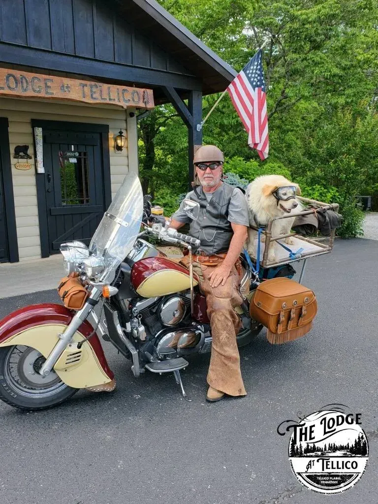 Man on motorcycle parked in front of a lodge, American flag. He's wearing a helmet and leather chaps.