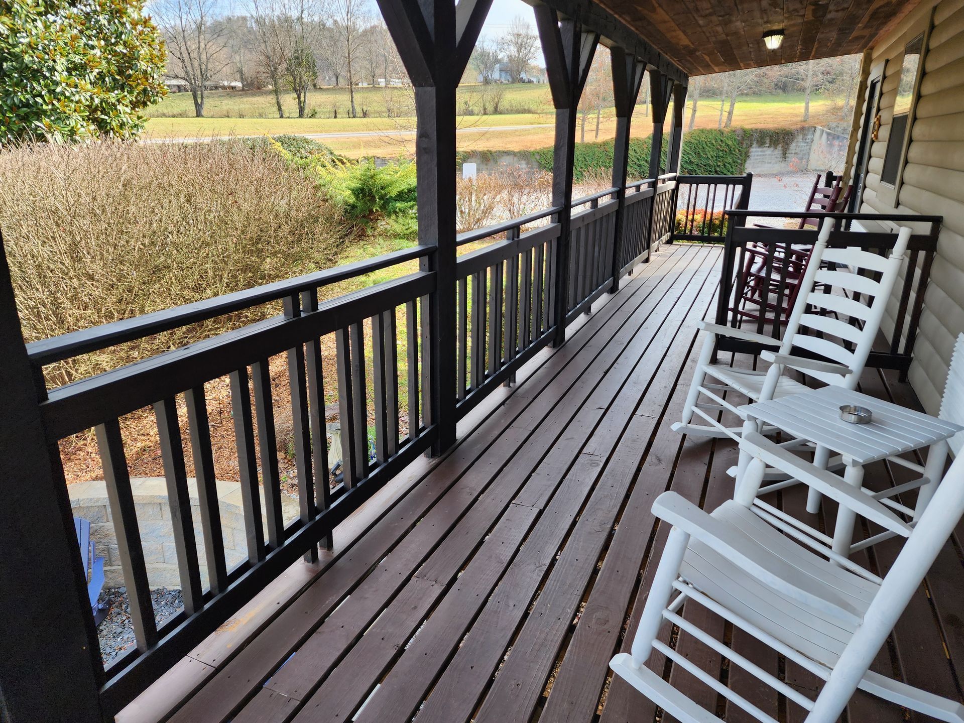 Covered porch with rocking chairs overlooking a green field and trees. Brown railing, wooden deck, and dark roof.
