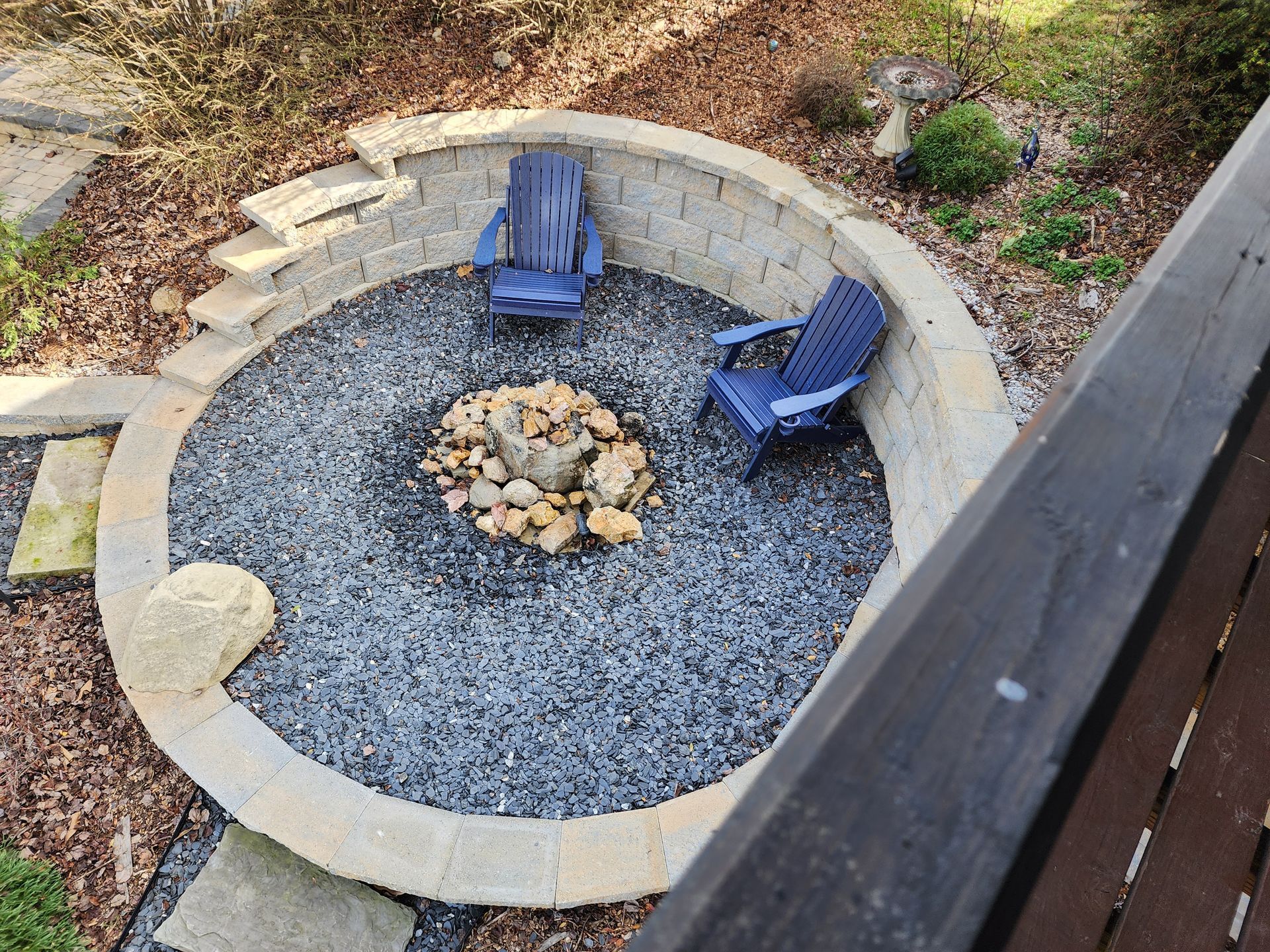 Circular fire pit area with two blue chairs on gravel, enclosed by a stone wall.
