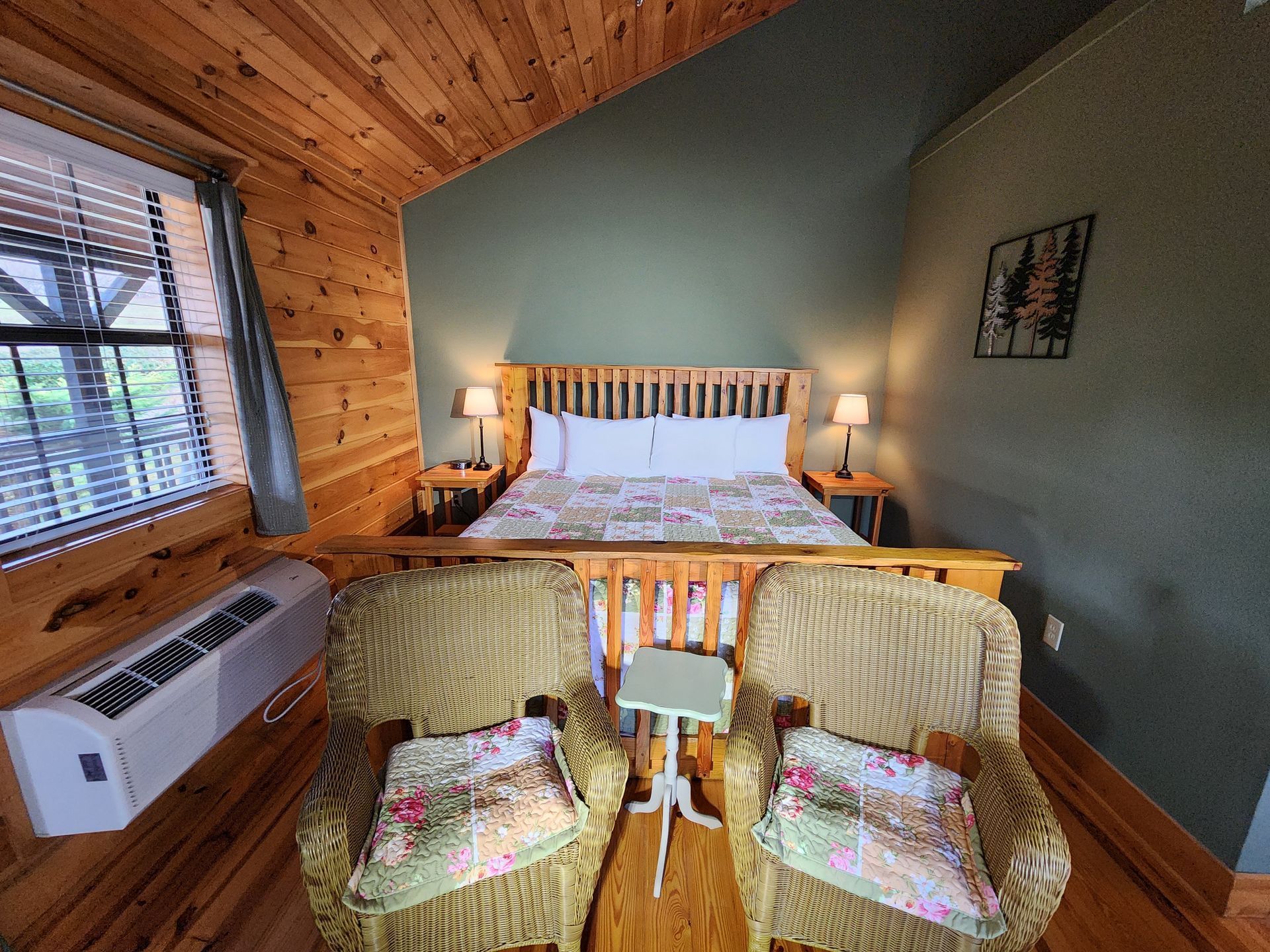 Cozy bedroom with wooden bed, wicker chairs, and small side table. Light-colored walls and wood panel ceiling.