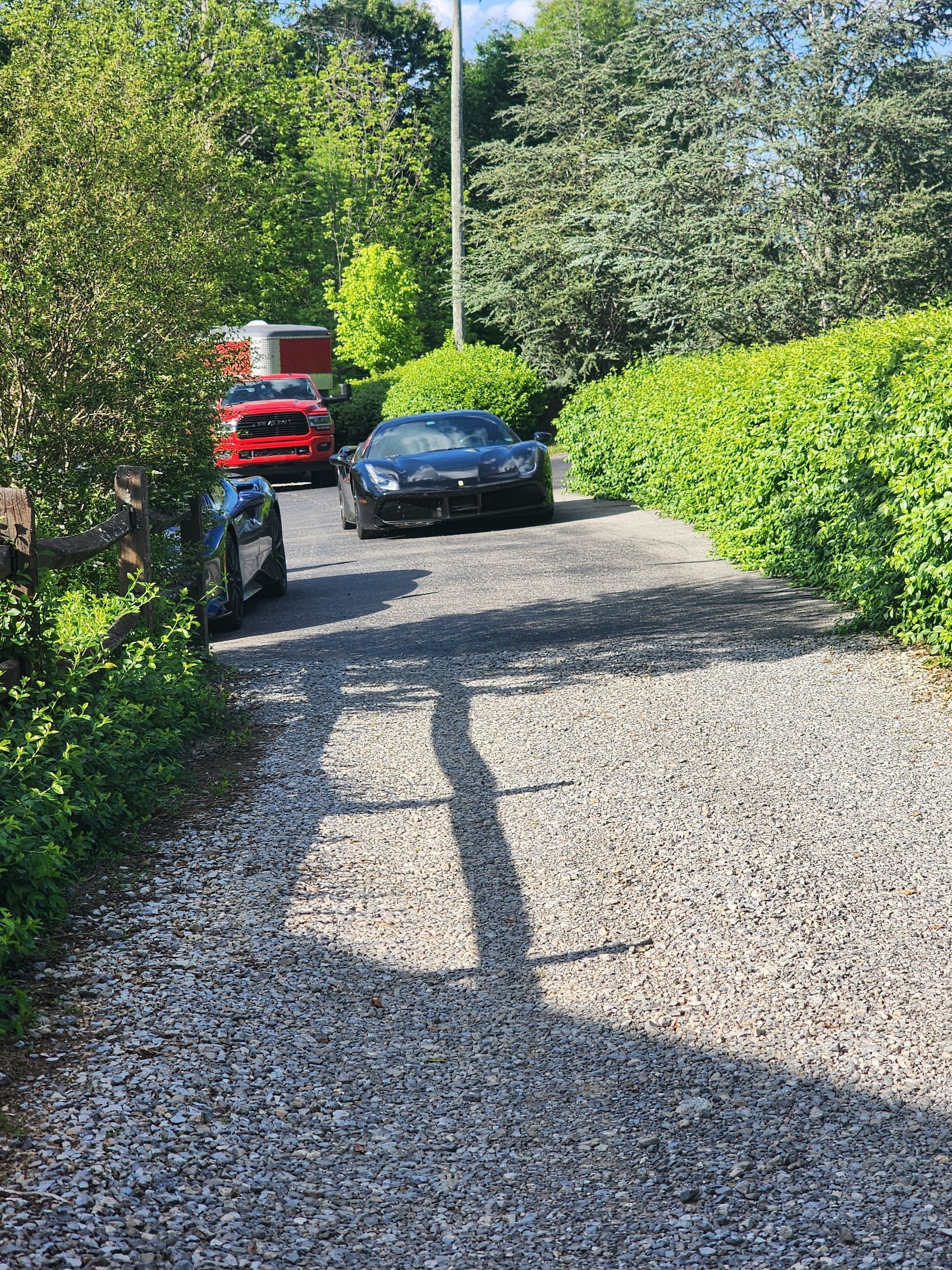 Black sports car on a gravel driveway, with a red truck in the background. Green bushes and trees surround.