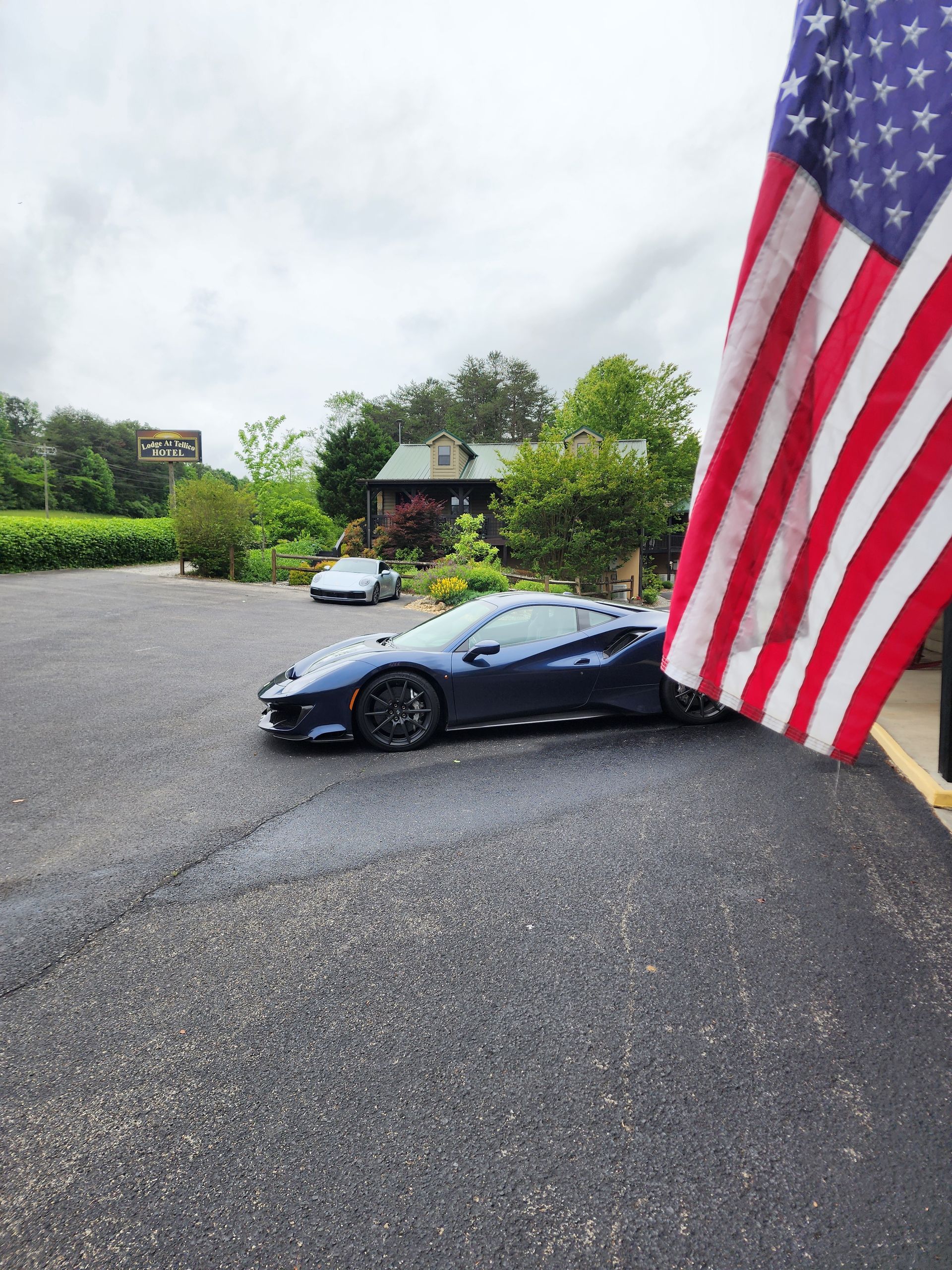 Blue sports car parked on asphalt with American flag in foreground, small building in background.