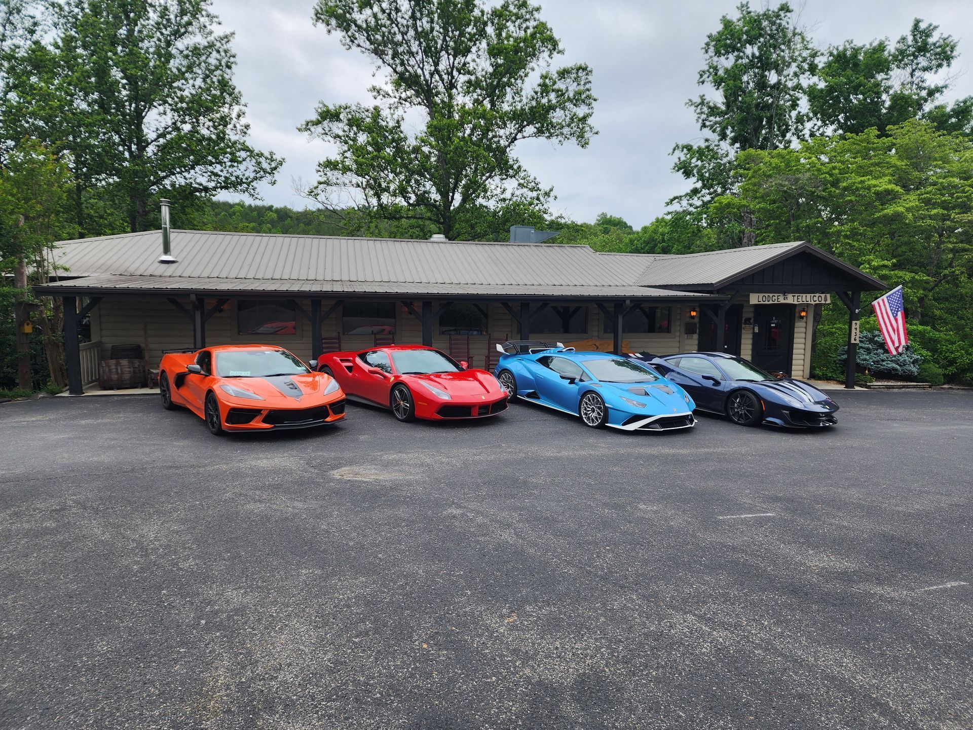 Four sports cars parked in front of a rustic building. Orange, red, light blue, and dark blue vehicles. Cloudy sky.