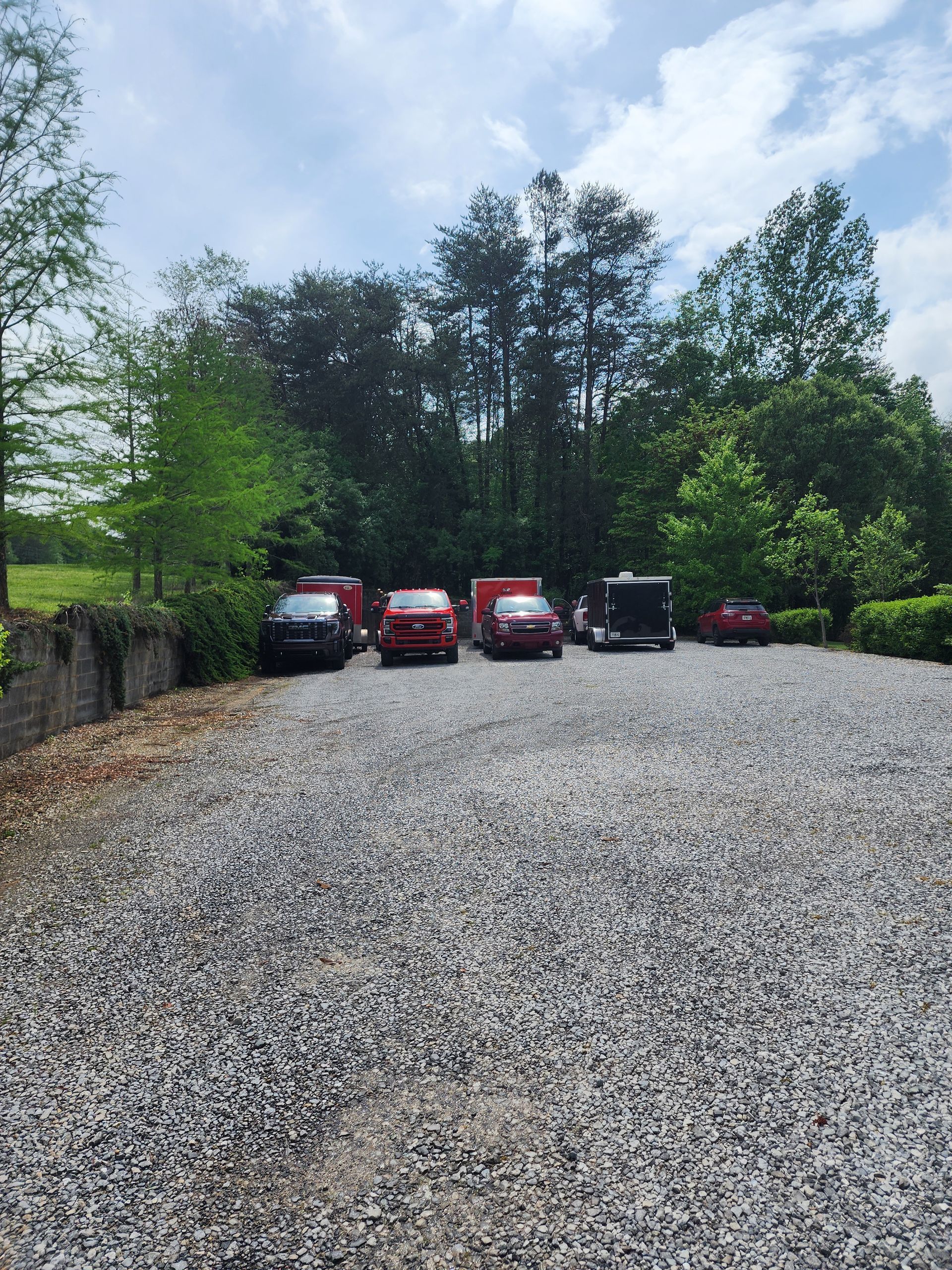 Gravel parking area with five vehicles in front of a tree line on a sunny day.