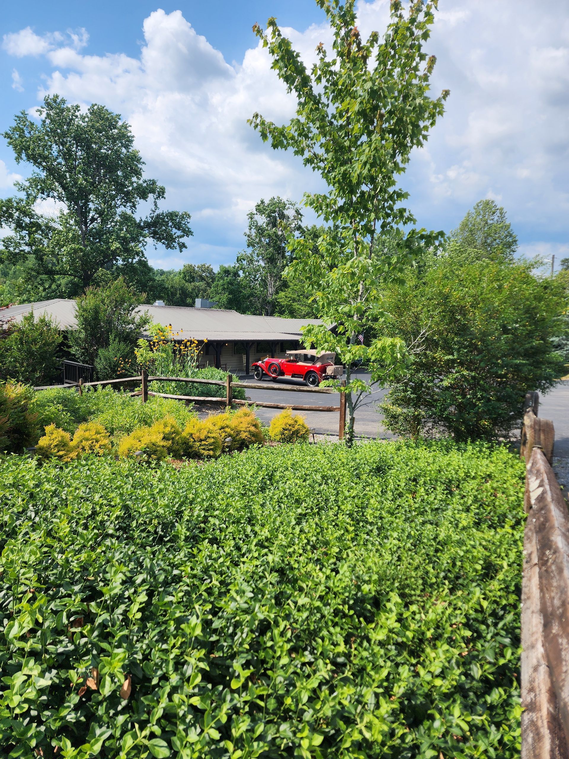 Red car parked outside a low building surrounded by lush greenery under a partly cloudy sky.