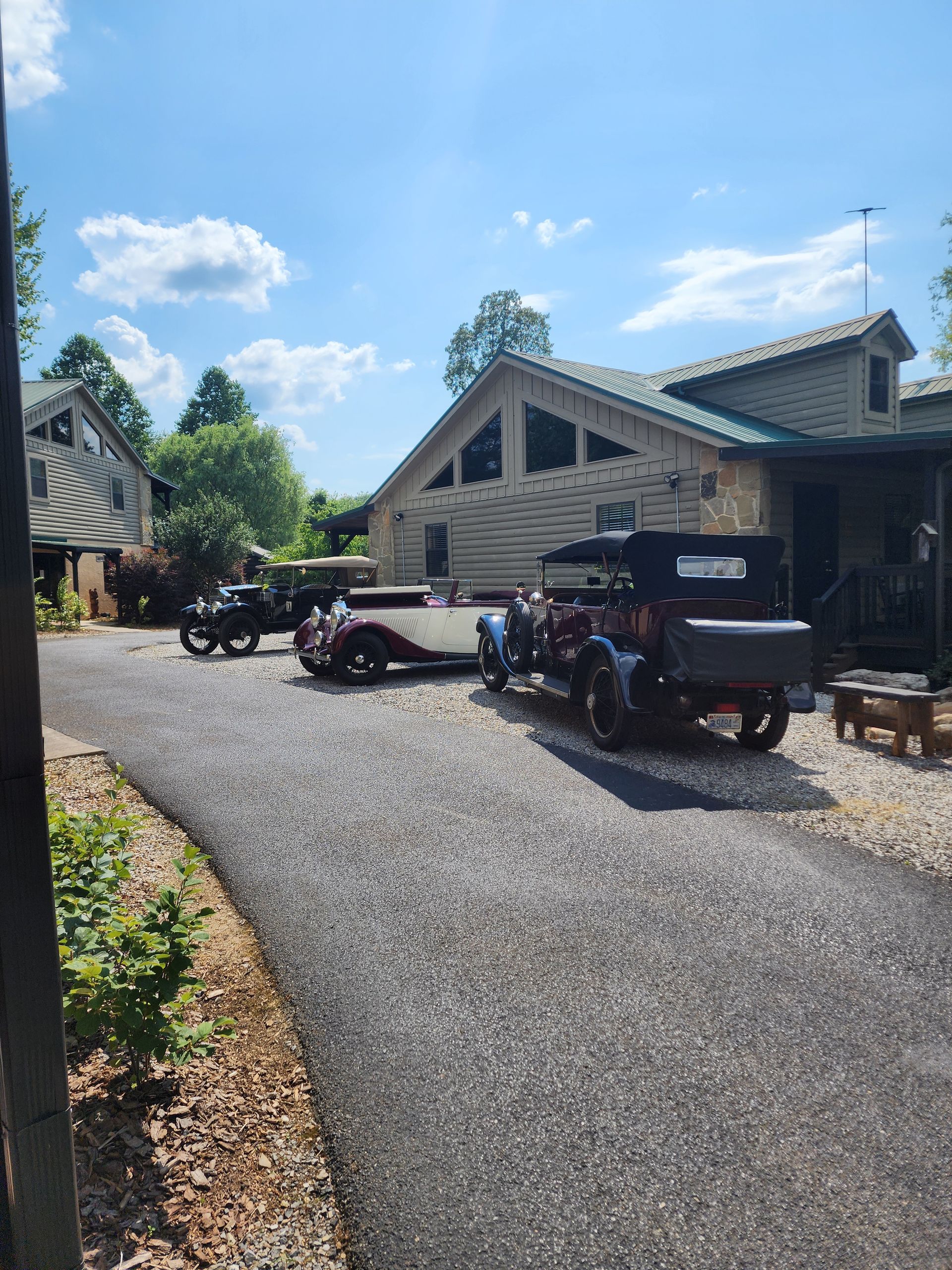 Vintage cars parked on gravel driveway in front of a wooden building on a sunny day.