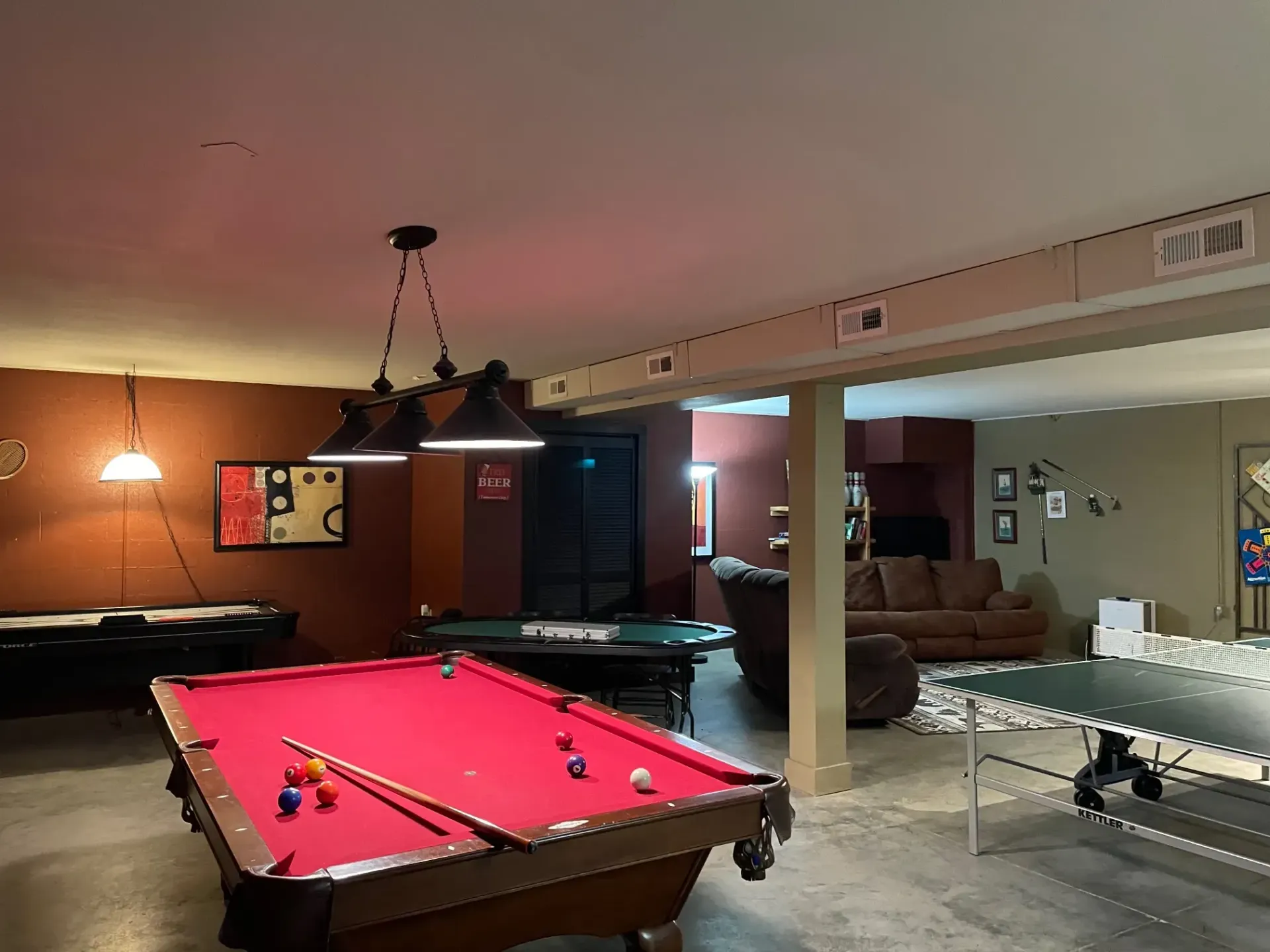 A basement with a red pool table, a ping pong table, and a brown sofa.