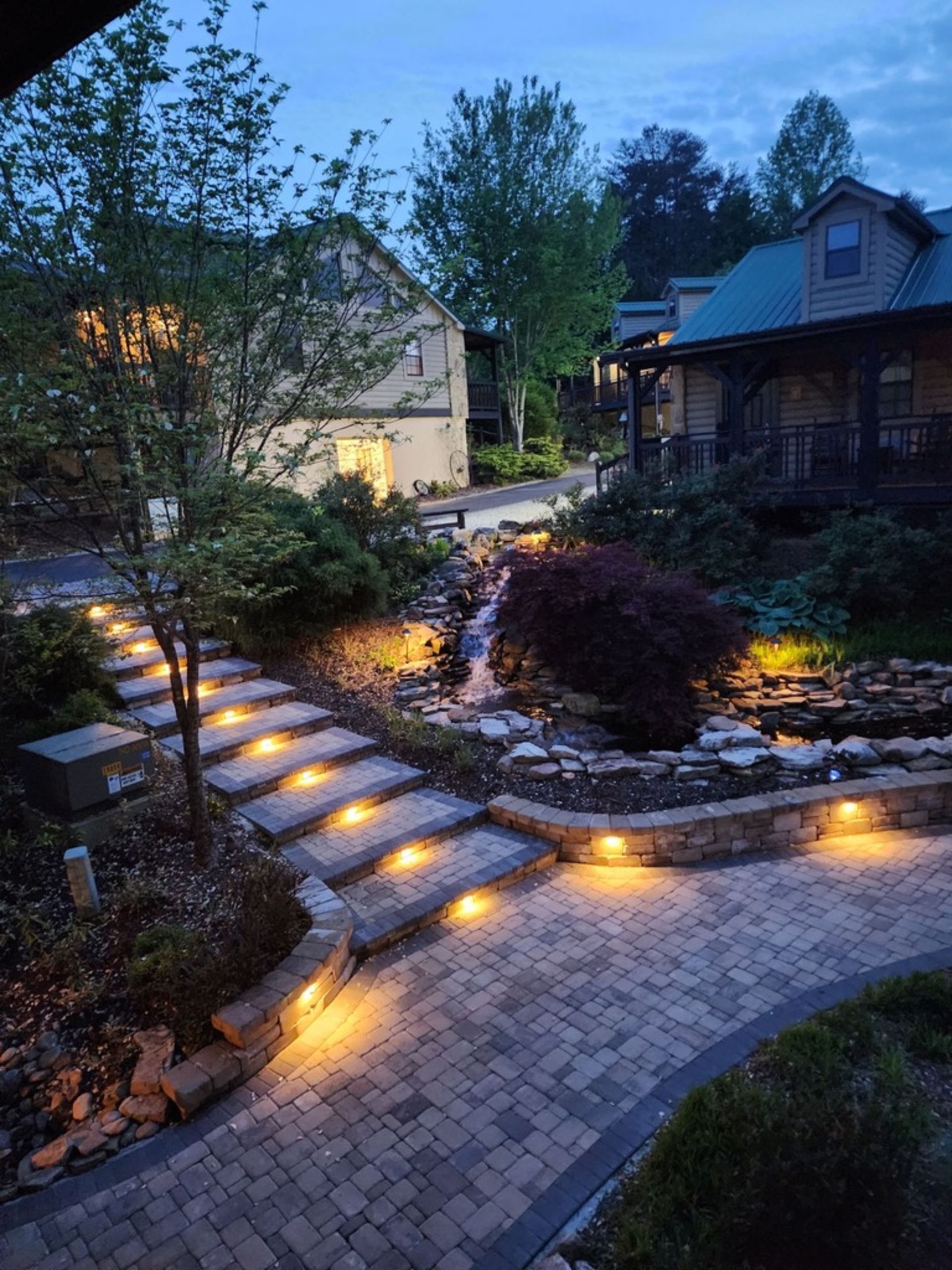 Stone pathway with steps, waterfall, and lit landscaping leading to buildings at dusk.