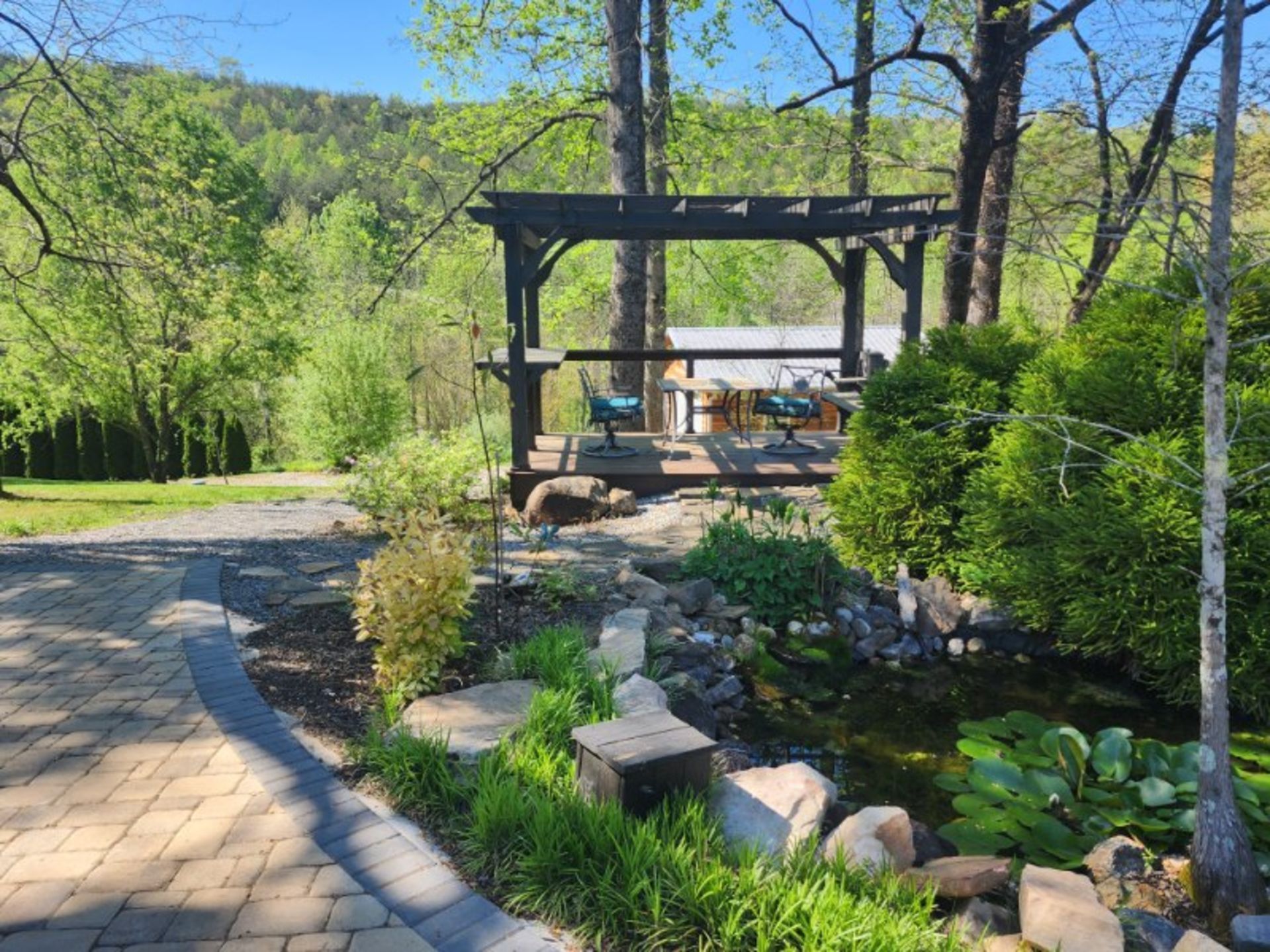 Patio with pergola overlooking a pond, surrounded by trees and greenery.