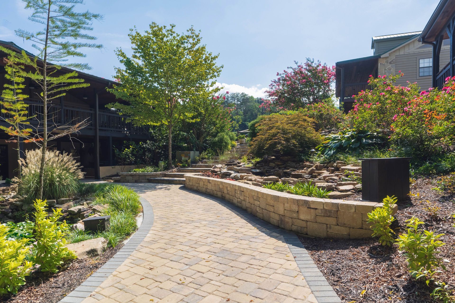 Stone pathway winds through landscaped garden with trees, flowers, and low walls under a sunny sky.