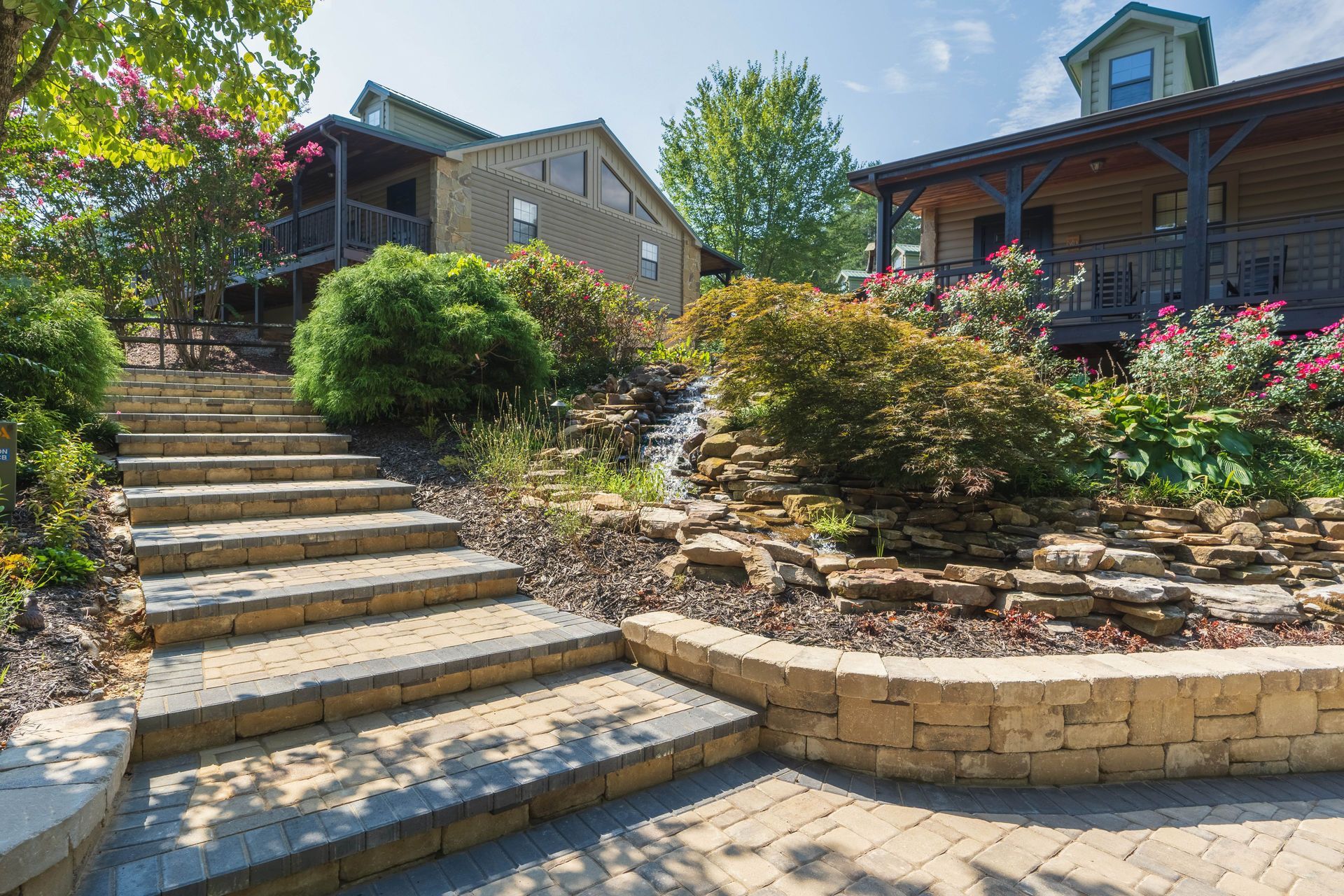 Stone steps lead up to a building with wooden siding, lush landscaping, and a small waterfall.