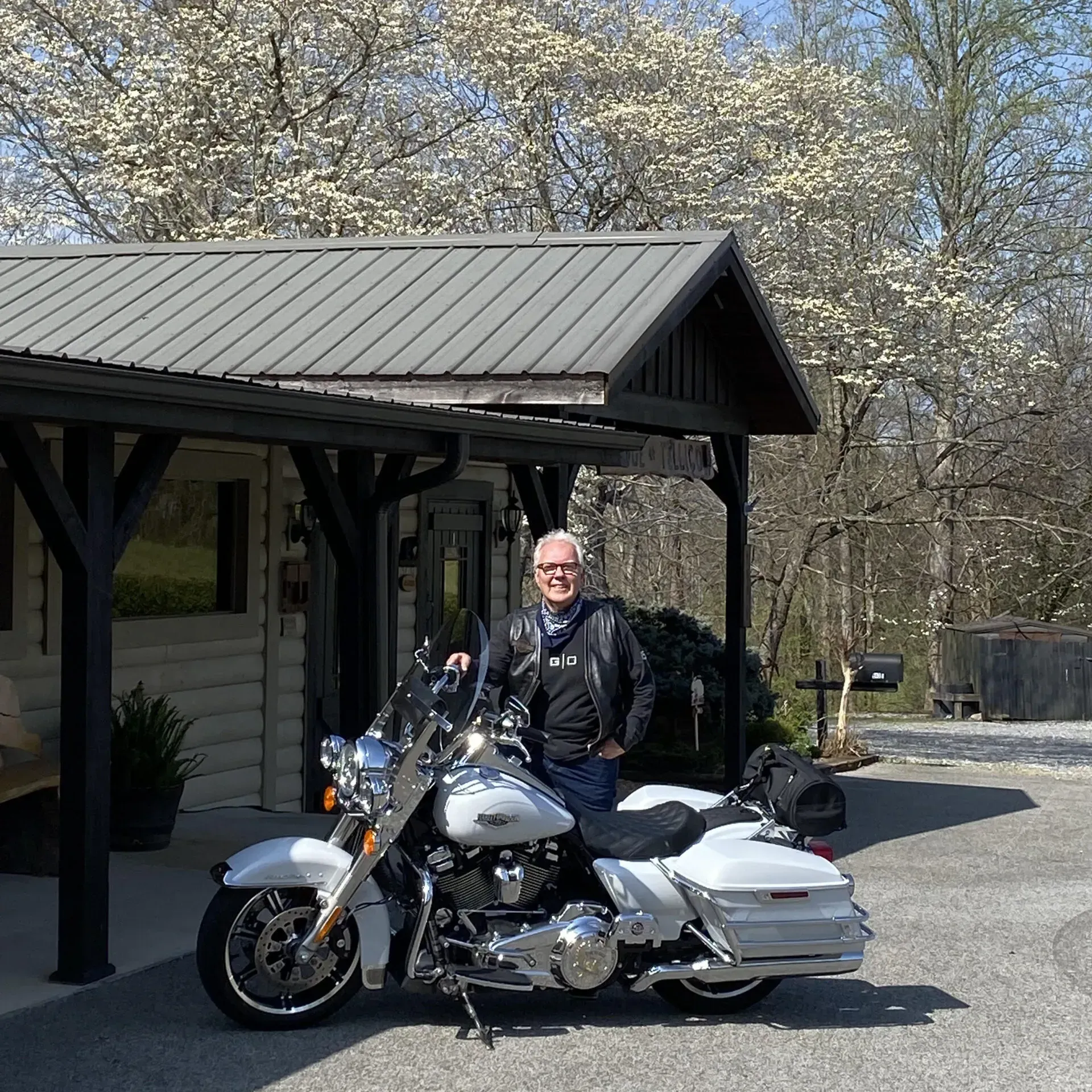 Man in black leather jacket poses next to a white motorcycle in front of a building with a gray roof.
