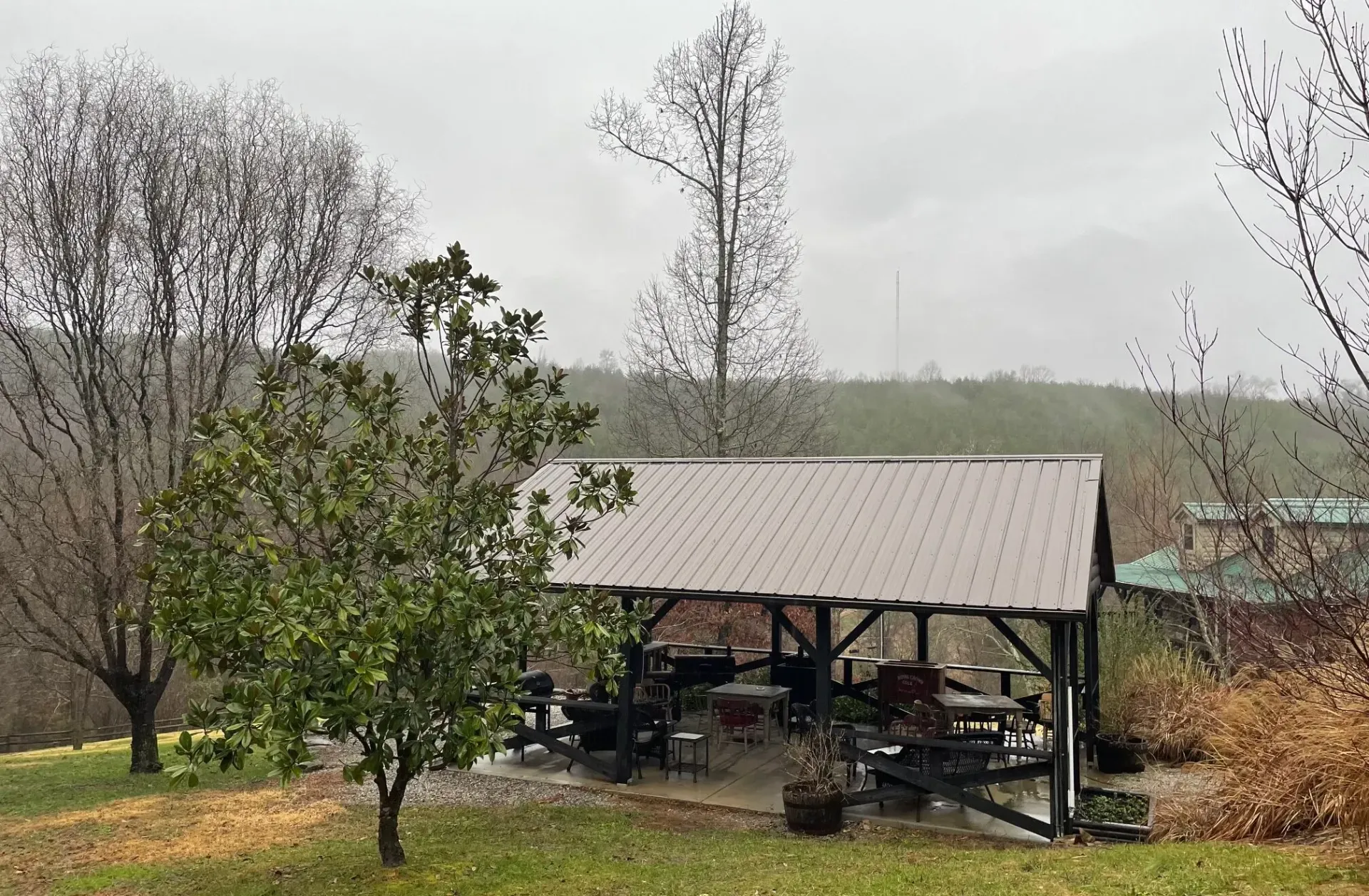 Covered outdoor seating area with tables and chairs, surrounded by trees and a hillside on a cloudy day.