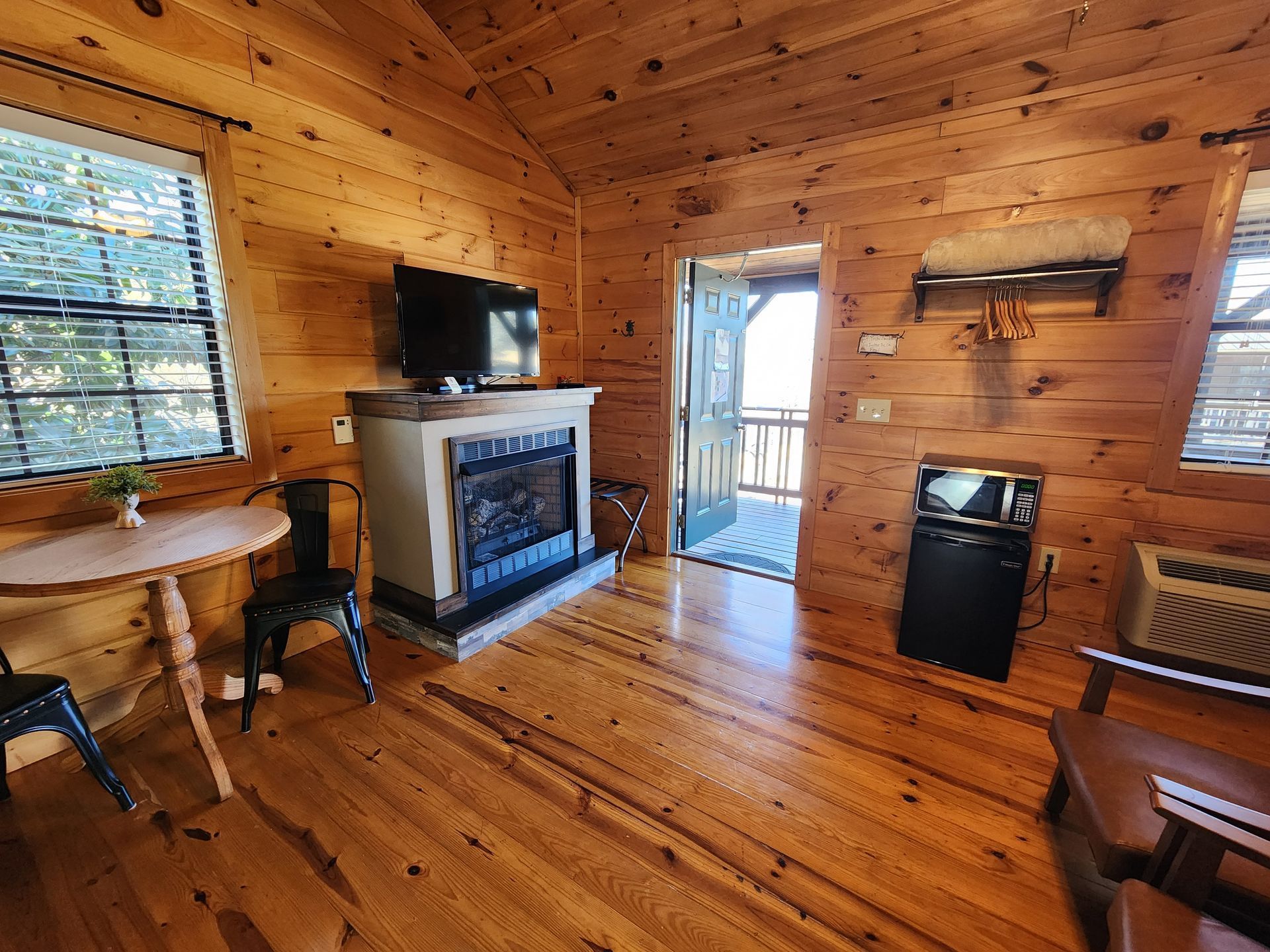 Cozy cabin interior with fireplace, TV, small table with chairs, microwave, and open door to balcony.