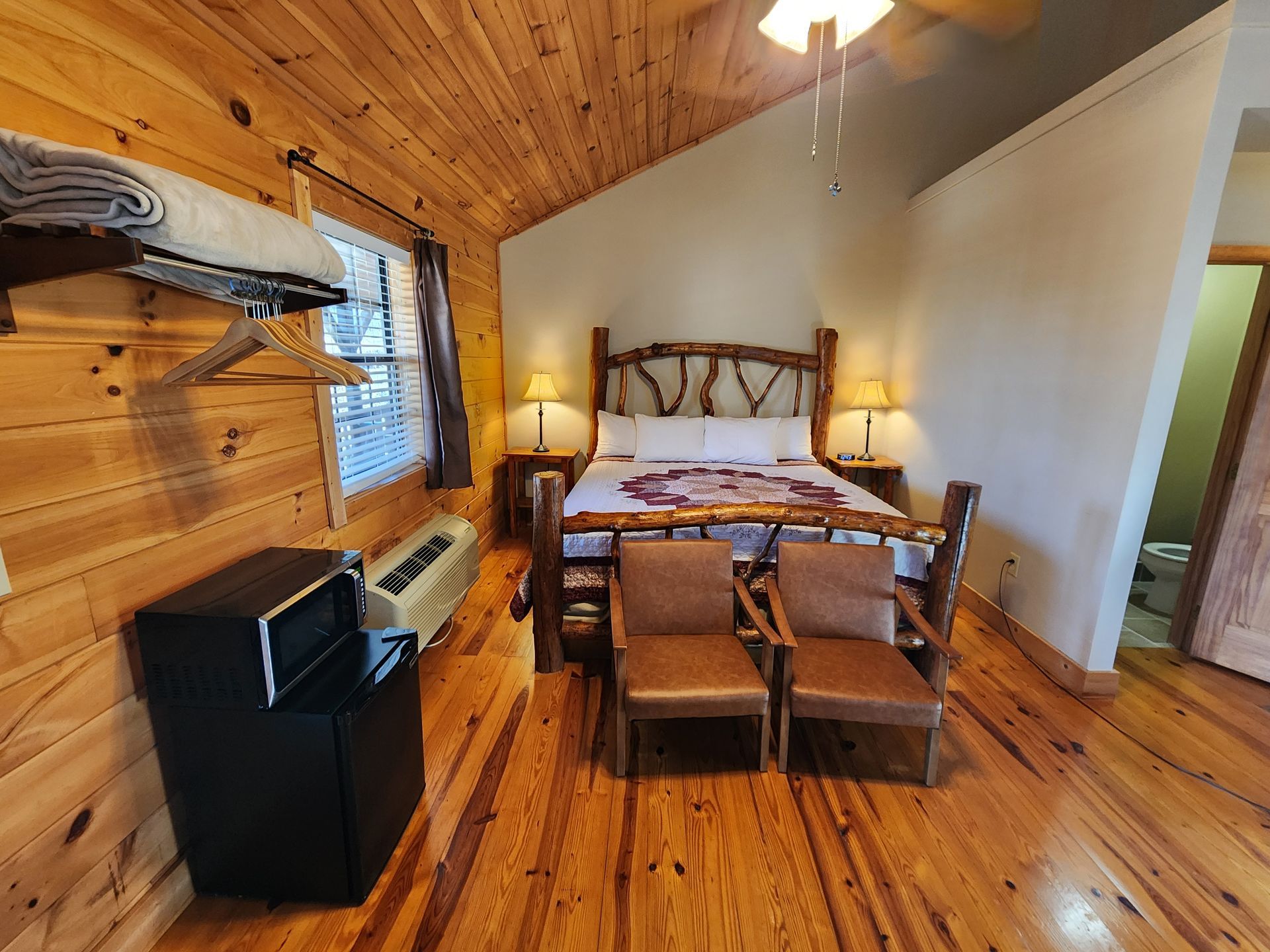 Cozy cabin bedroom with wooden walls and ceiling, a bed, chairs, and a microwave.