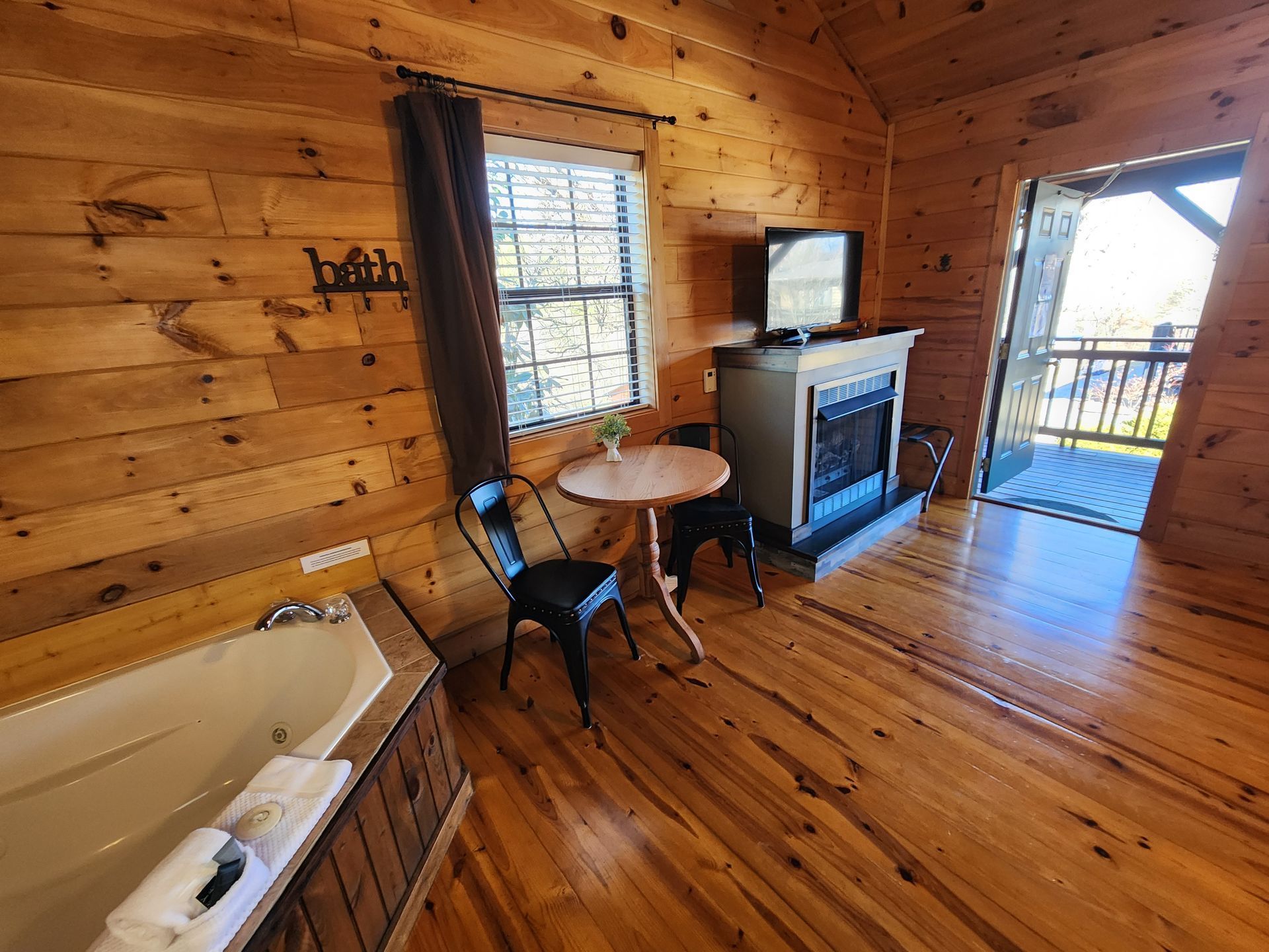 Cozy cabin interior with a fireplace, jacuzzi tub, table, chairs, and open doorway to a balcony.