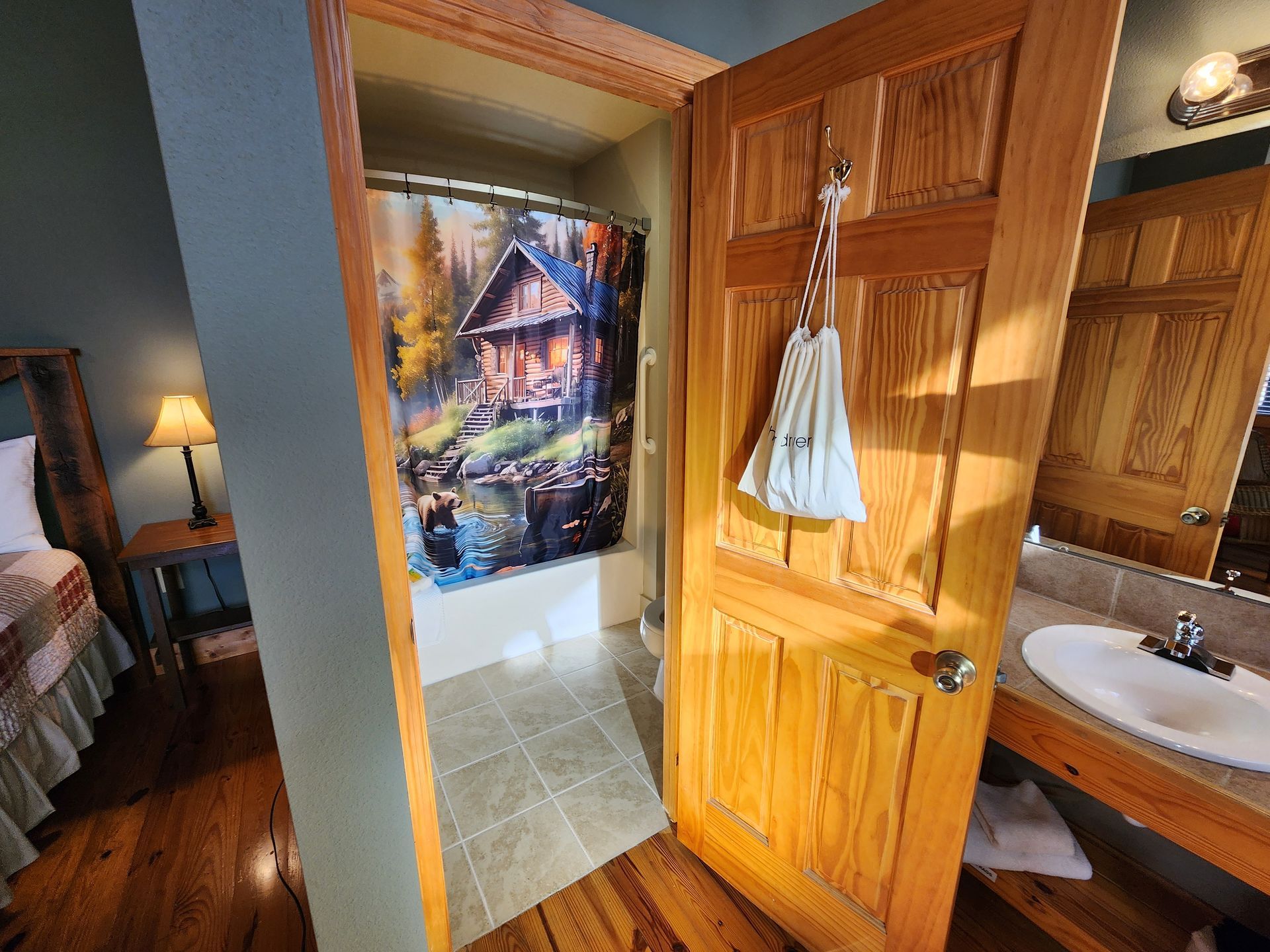 Open doorway to a bathroom with a shower curtain depicting a cabin scene. A sink and wooden door are visible.