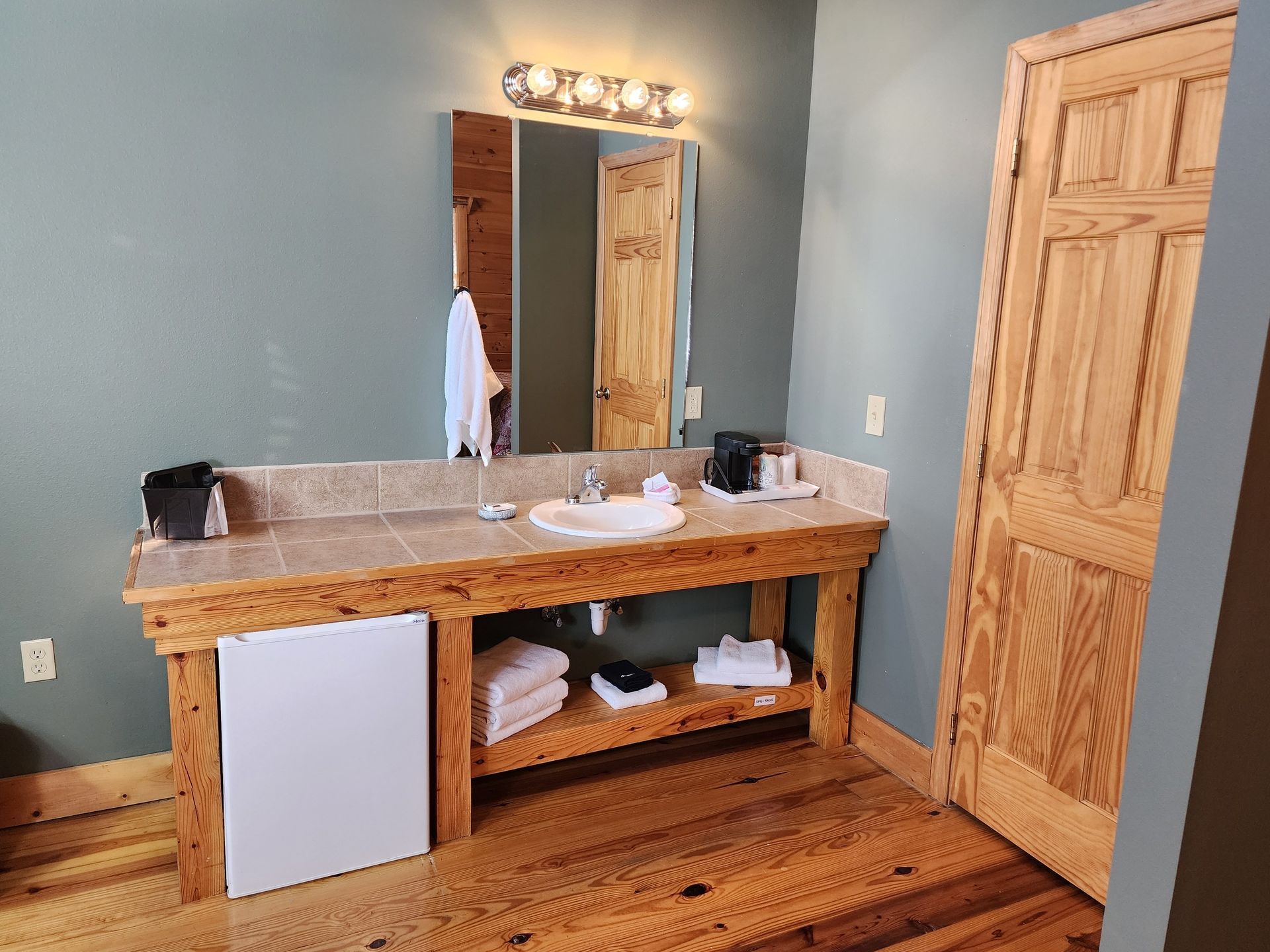 Bathroom with wooden vanity, sink, mirror, mini-fridge, towels, and a closed wooden door.