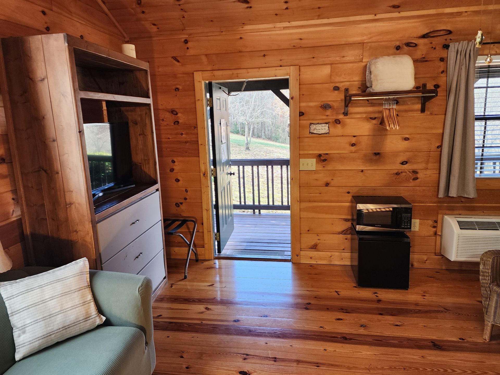 Interior of a wooden cabin with open door to a small porch. Includes a TV cabinet, couch, and microwave.