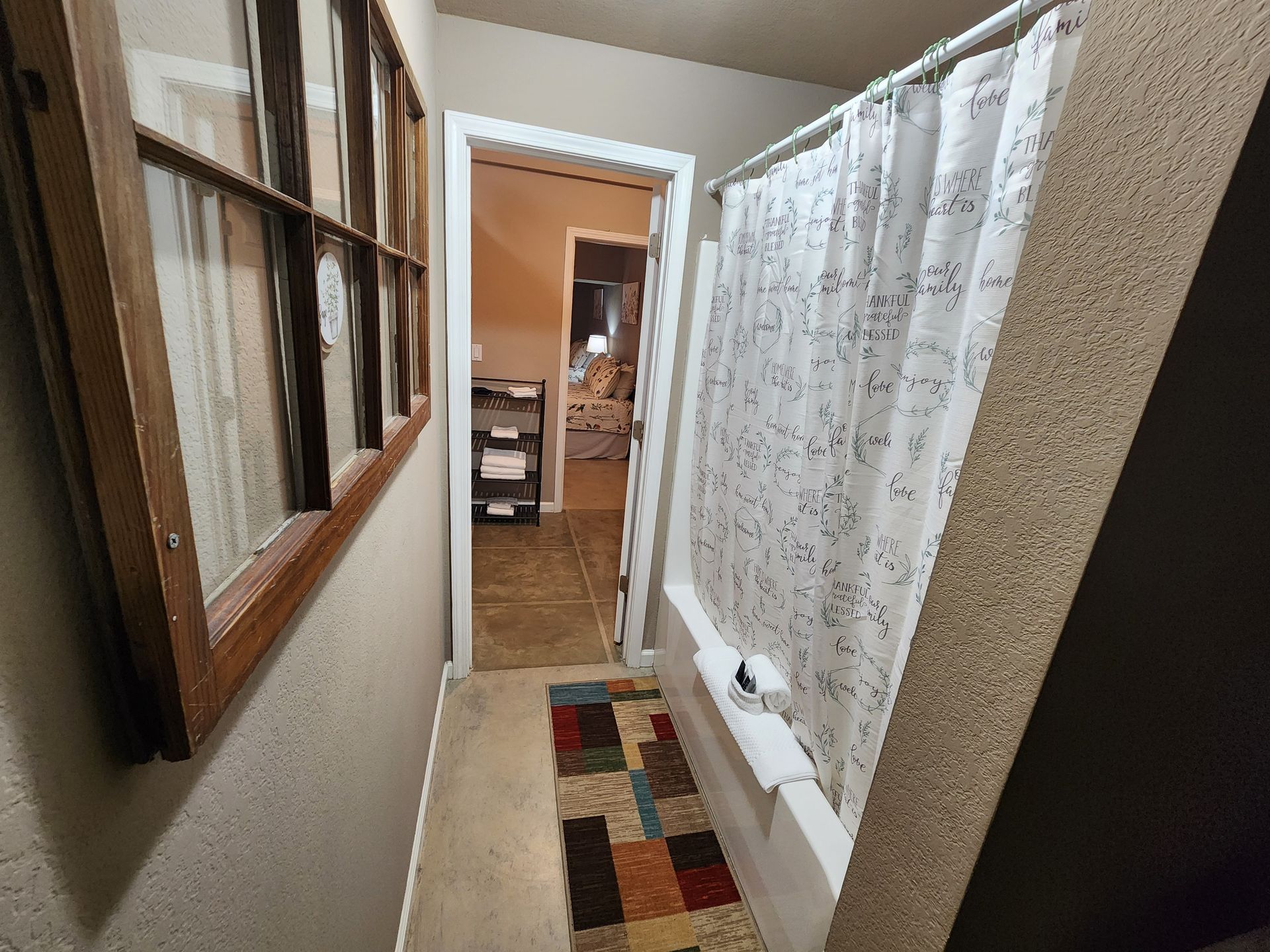 Narrow bathroom with shower, window, open doorway to bedroom, and colorful rug.