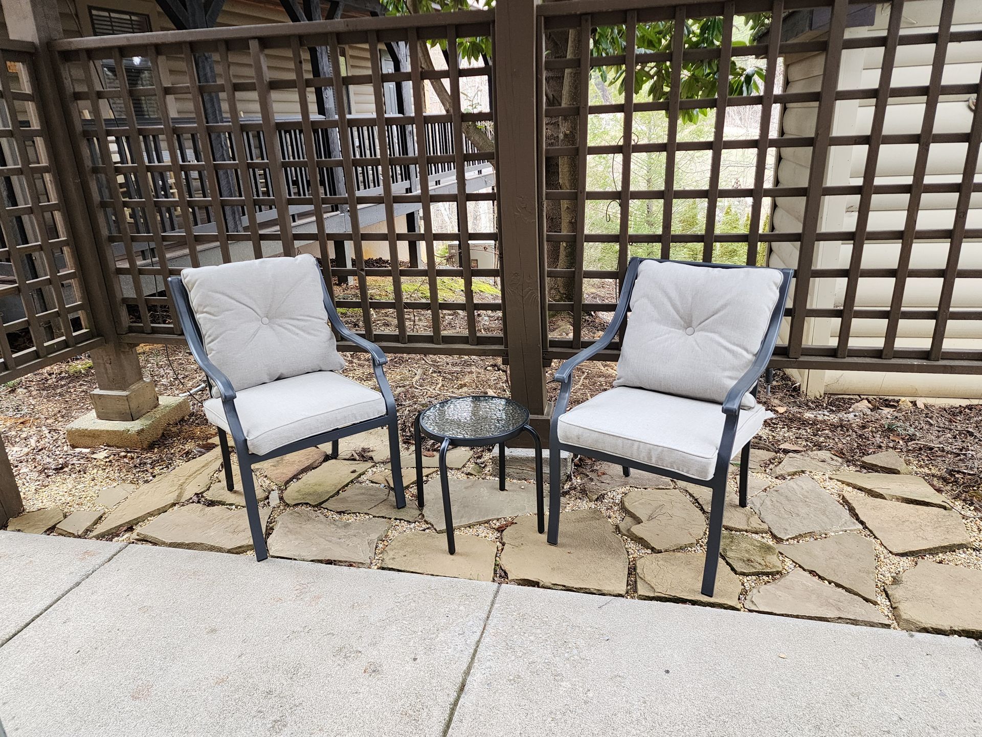 Two cushioned chairs face each other with a small table in between on a stone patio, enclosed by a lattice fence.