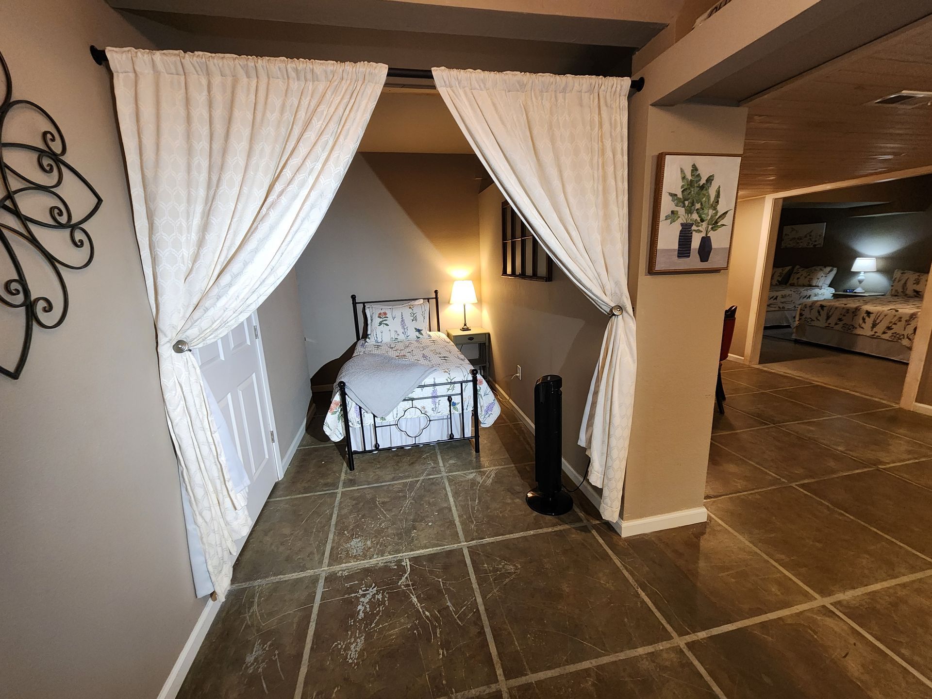 A small bed alcove with white curtains, a nightstand, and an open doorway to other rooms.