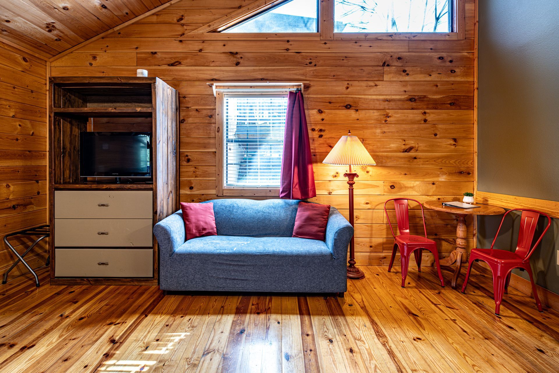 Cozy cabin interior with wood paneling, couch, TV, small table with red chairs, and window with purple curtain.