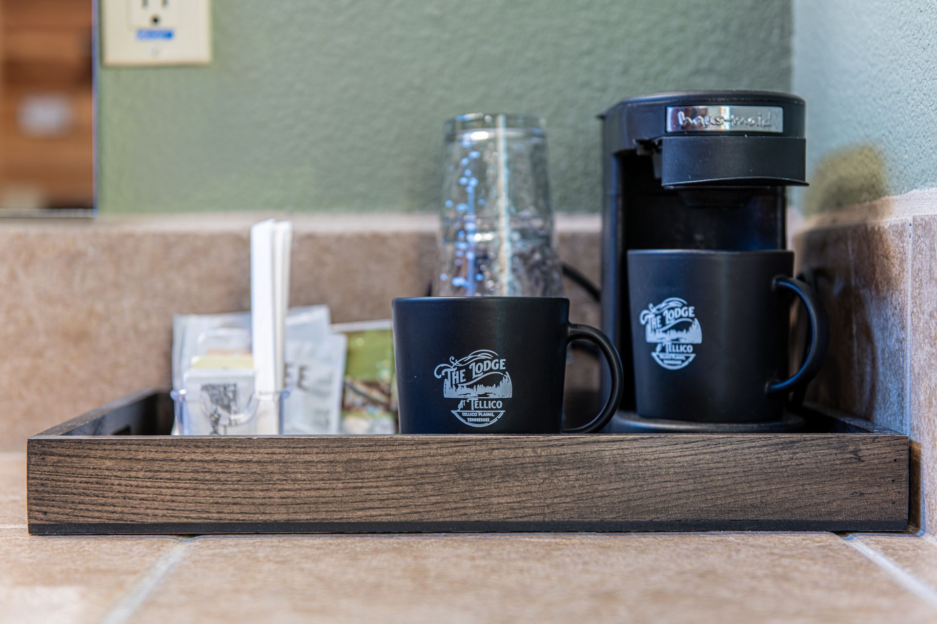 Coffee station in a bathroom: black coffee maker, mugs, and supplies on a wooden tray, beige countertop, and green wall.