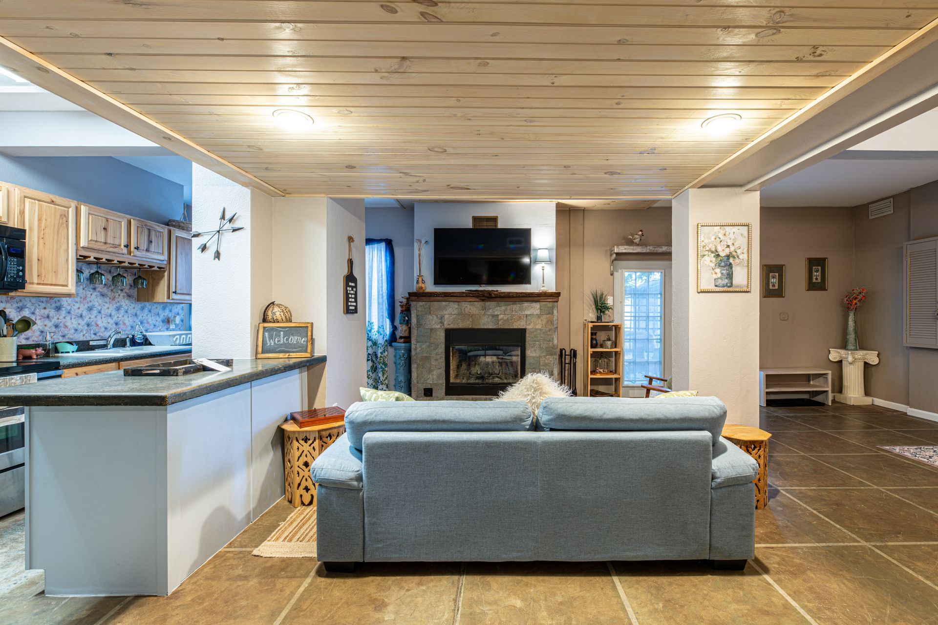 Living room with stone fireplace, open kitchen, blue couch, and wood ceiling.