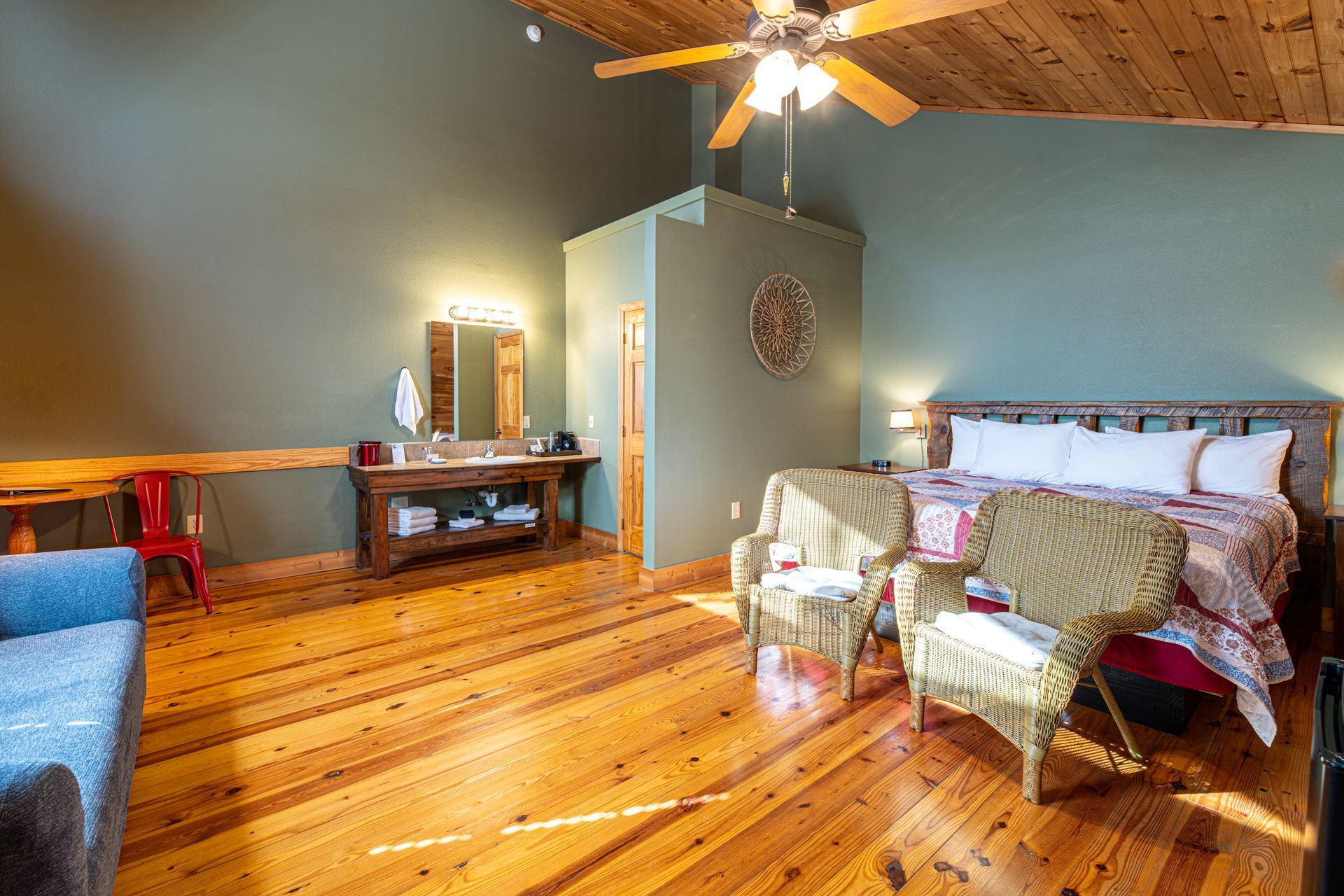 Bedroom with wood floors, ceiling fan, queen bed, two wicker chairs, and a vanity. Green walls.
