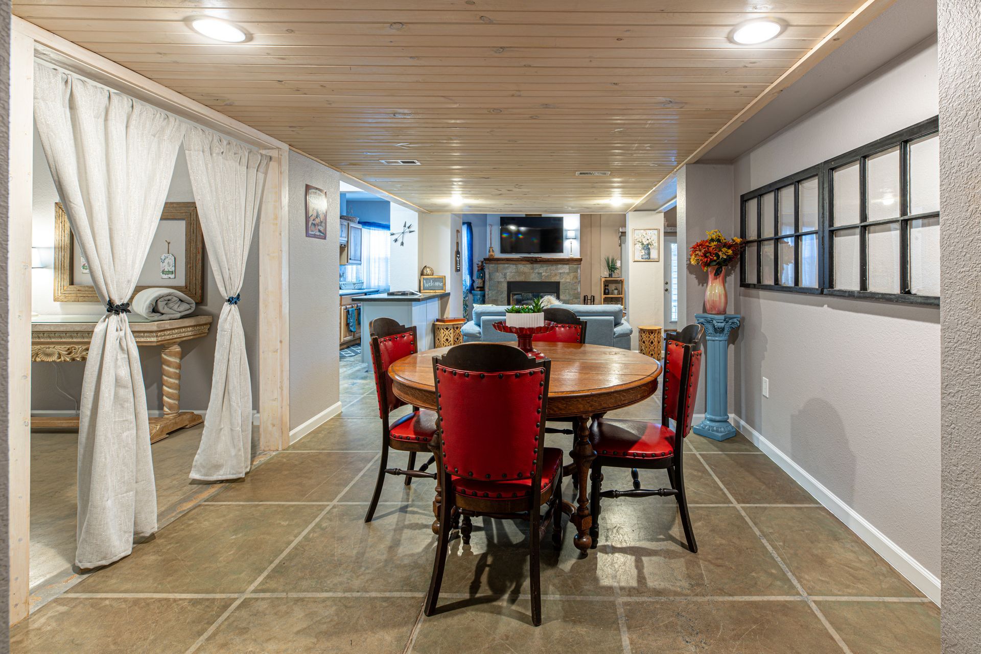 Dining area with a round wooden table, red chairs, and curtains to the left, leading to a living room.