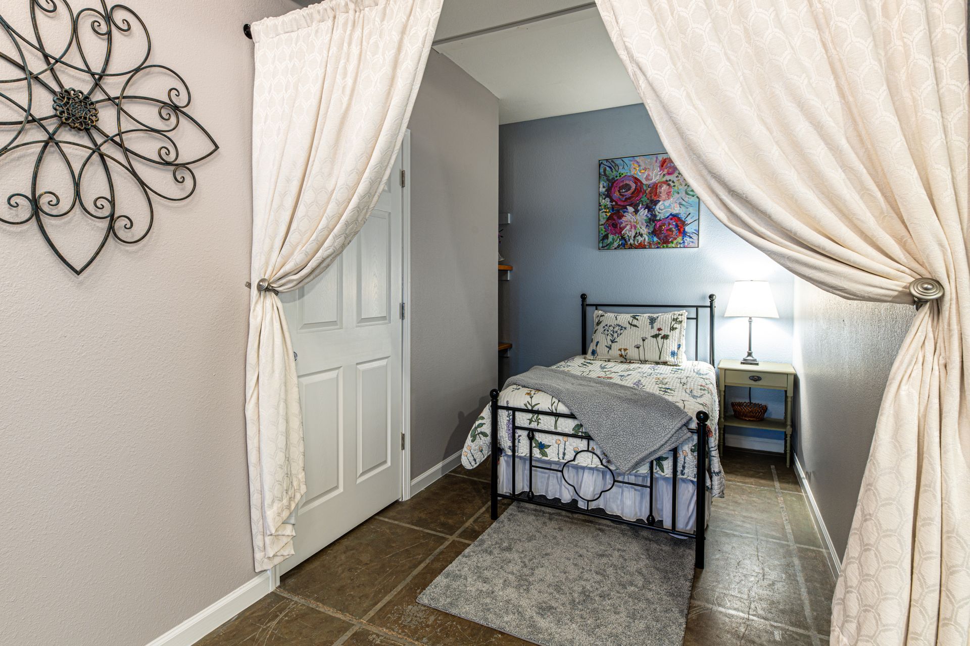 Small bedroom with curtain doorway, bed, nightstand, floral art, and a decorative wall hanging.