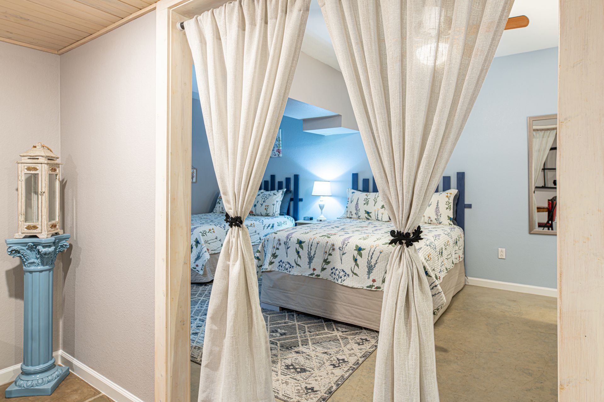 View of two twin beds through a doorway with sheer curtains, blue and beige tones.