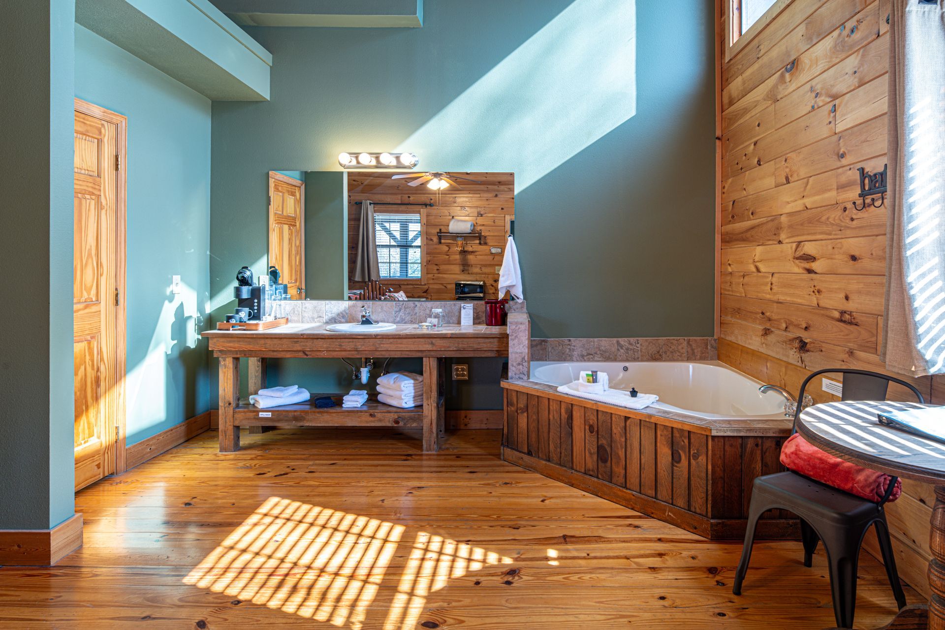 Bathroom with wood paneling, teal walls, a tub, large sink, and wood floors. Sunlight streams through a window.
