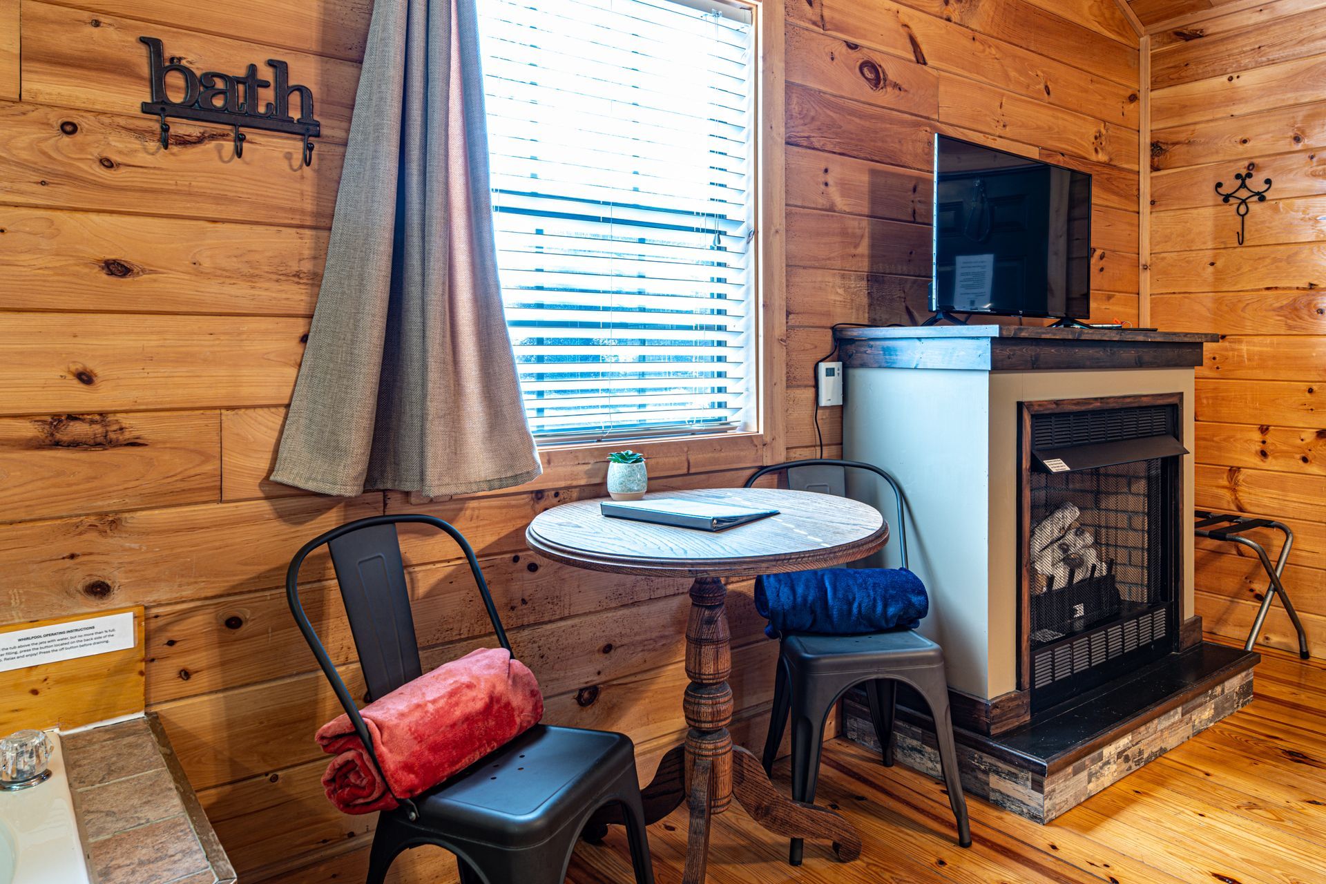 Cozy cabin interior: small round table, chairs, fireplace with TV, and window. Wooden walls.