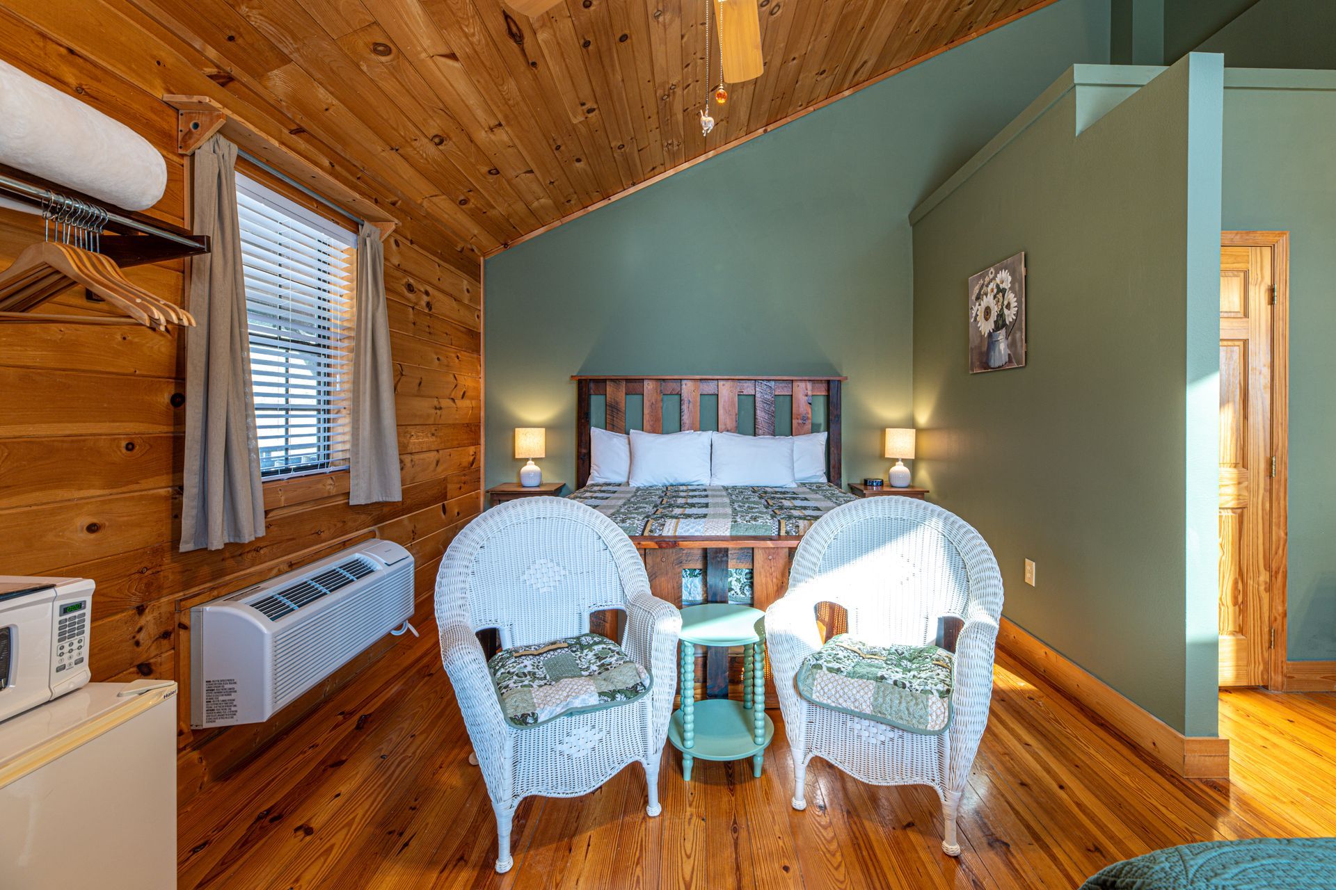 Bedroom with wooden walls and ceiling, queen bed, two white wicker chairs, and small teal side table.