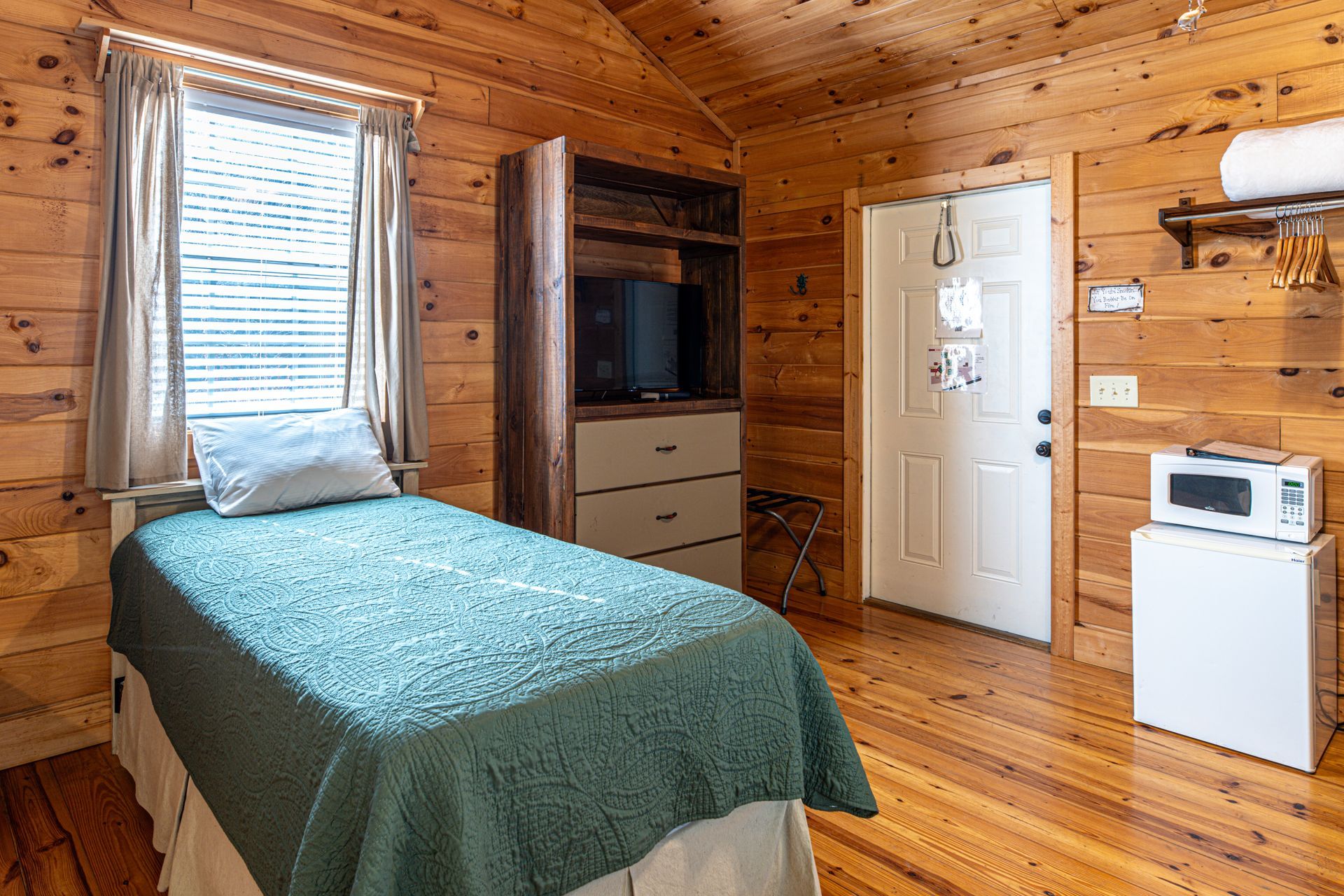 Cozy cabin room with wooden walls and floor, bed with green cover, window, door, and microwave.