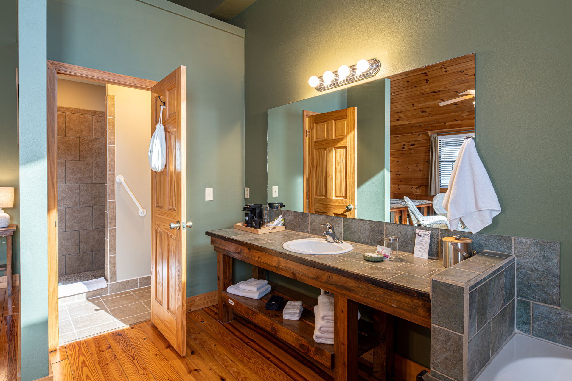 Bathroom with wooden vanity, large mirror, and open doorway to a shower. Green walls, wood flooring.