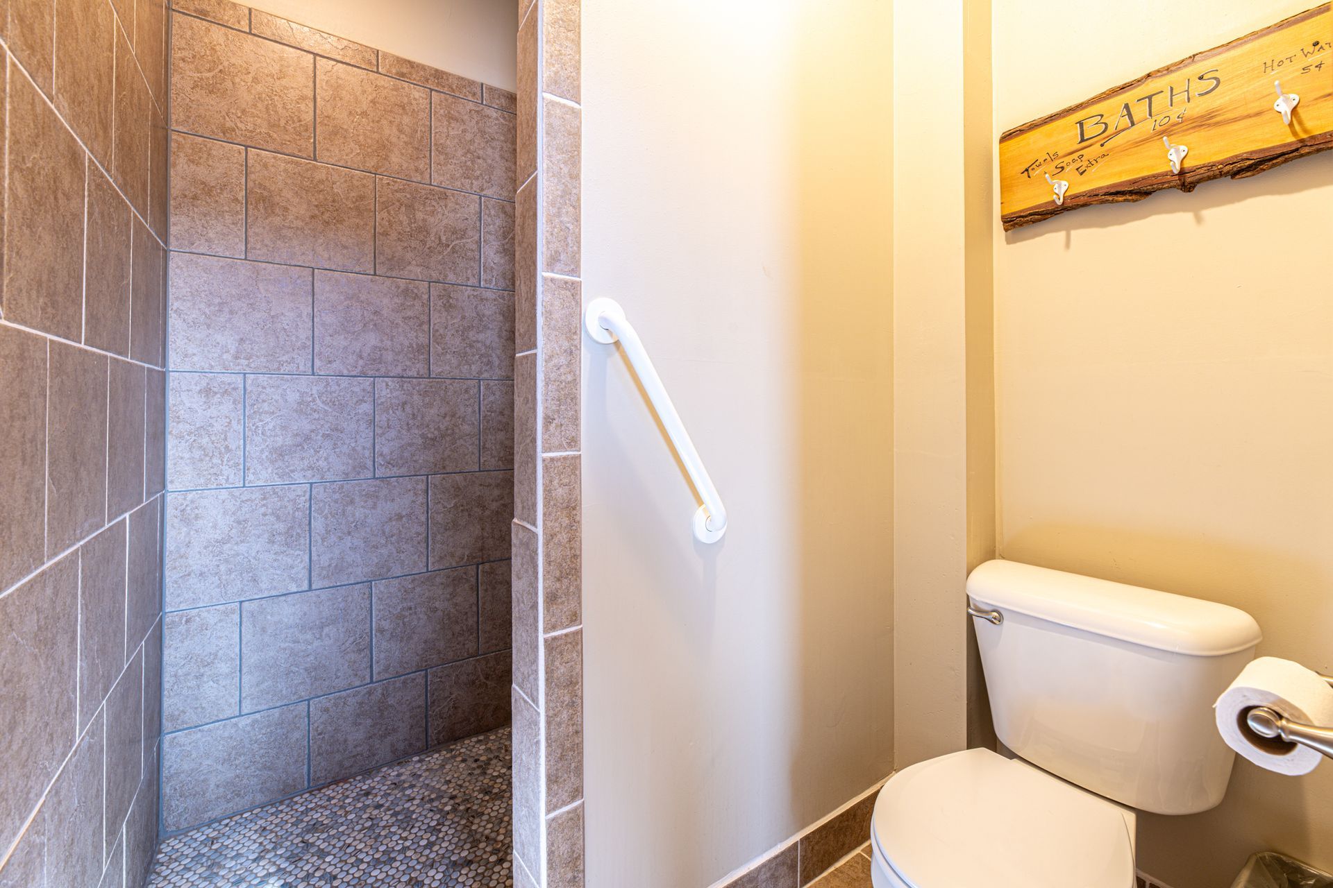 Bathroom with walk-in shower, toilet, and grab bar. Beige walls, stone shower, and a wooden sign.
