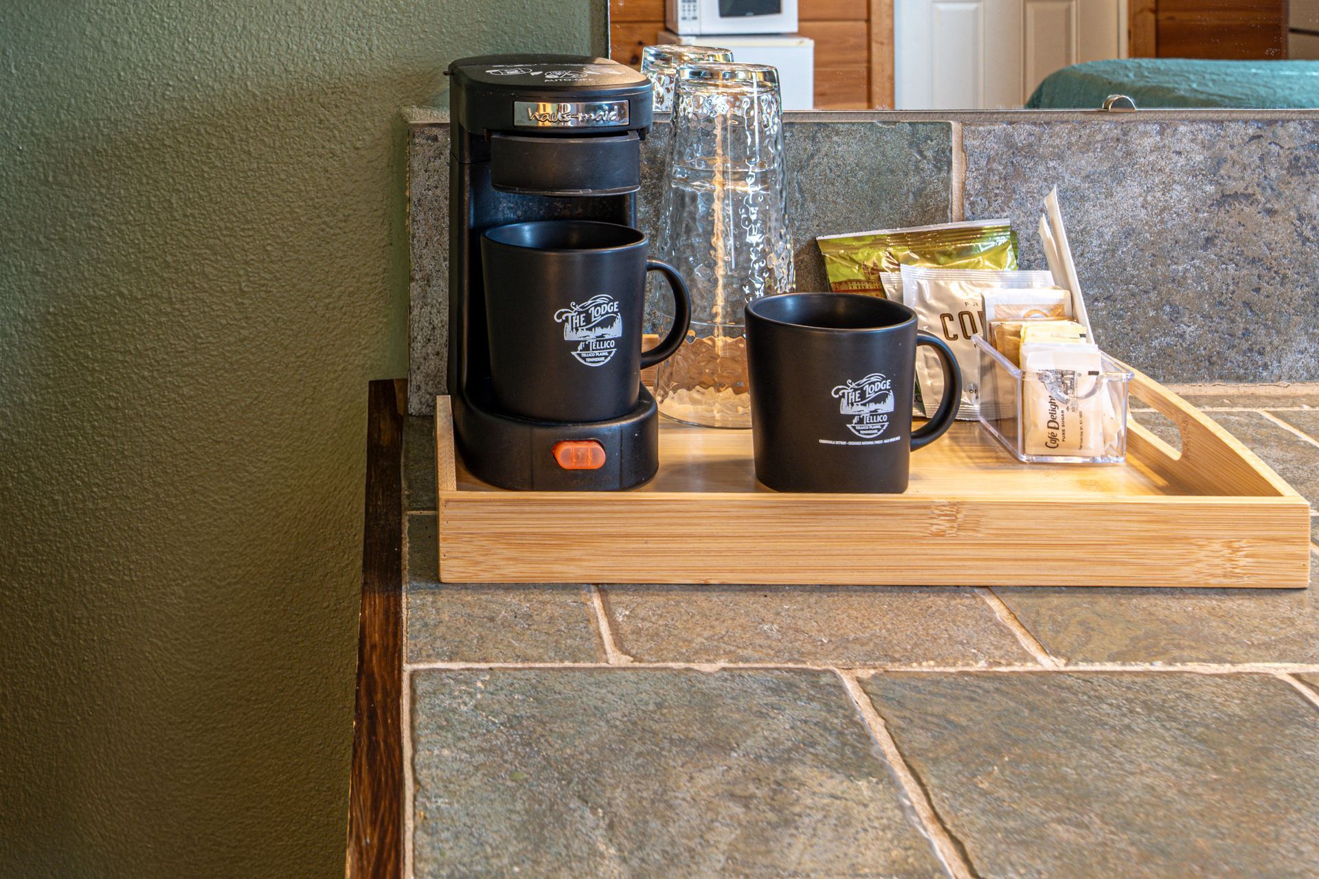 Coffee maker, mugs, and supplies on a wooden tray atop a slate countertop.