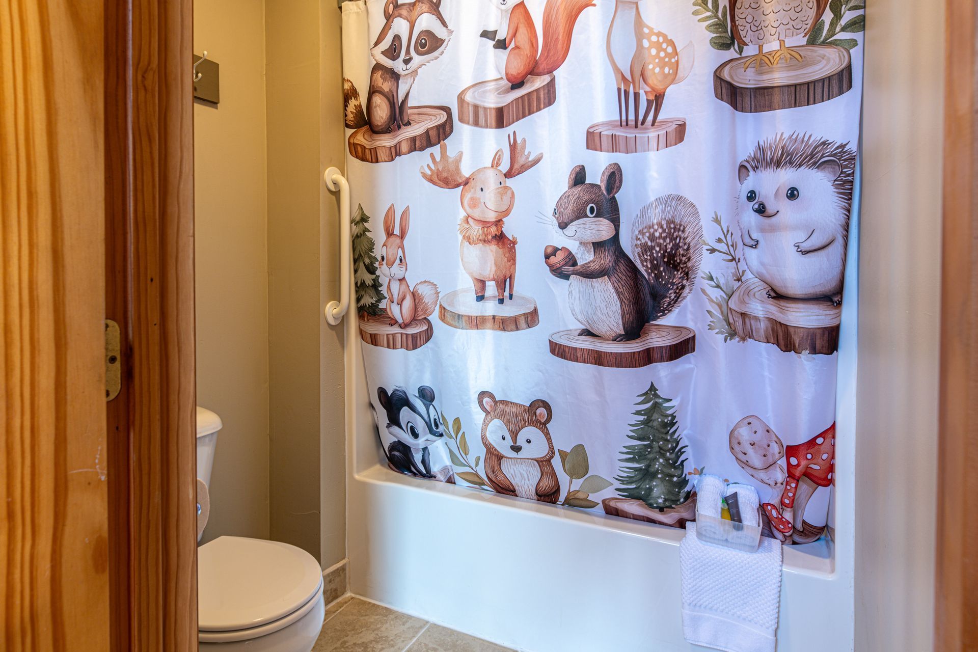Bathroom with woodland-themed shower curtain, white tub, and toilet. Light walls and wooden door.