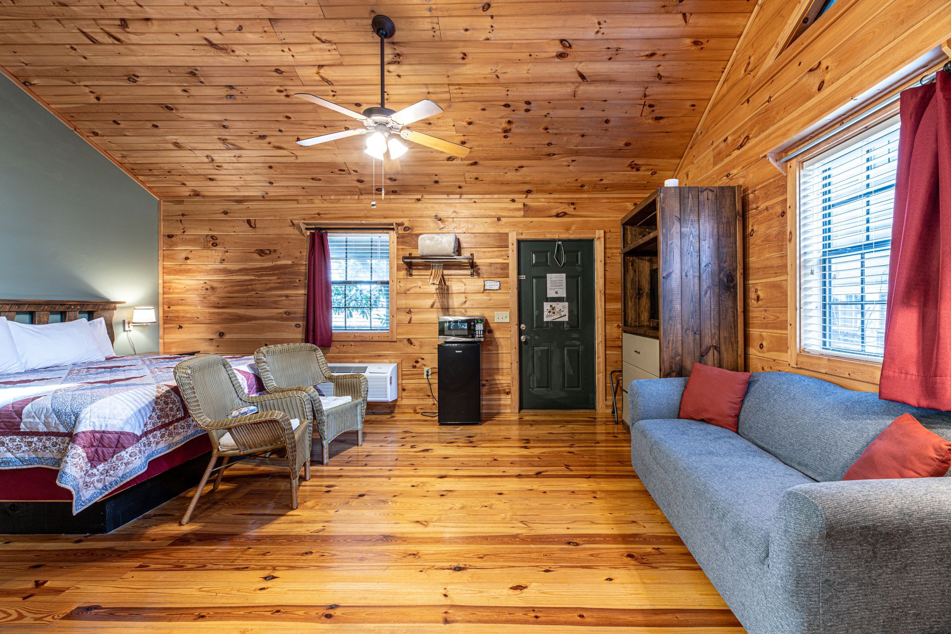 Cozy cabin interior: bed, sofa, chairs, wooden walls, ceiling fan, and windows.
