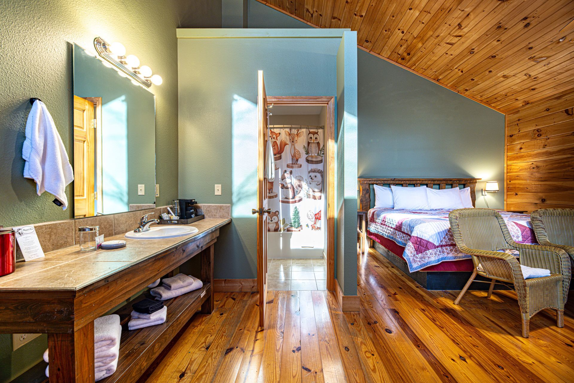 Hotel room: bathroom with sink, mirror, and toiletries, connected to a bedroom with bed and wicker chairs.