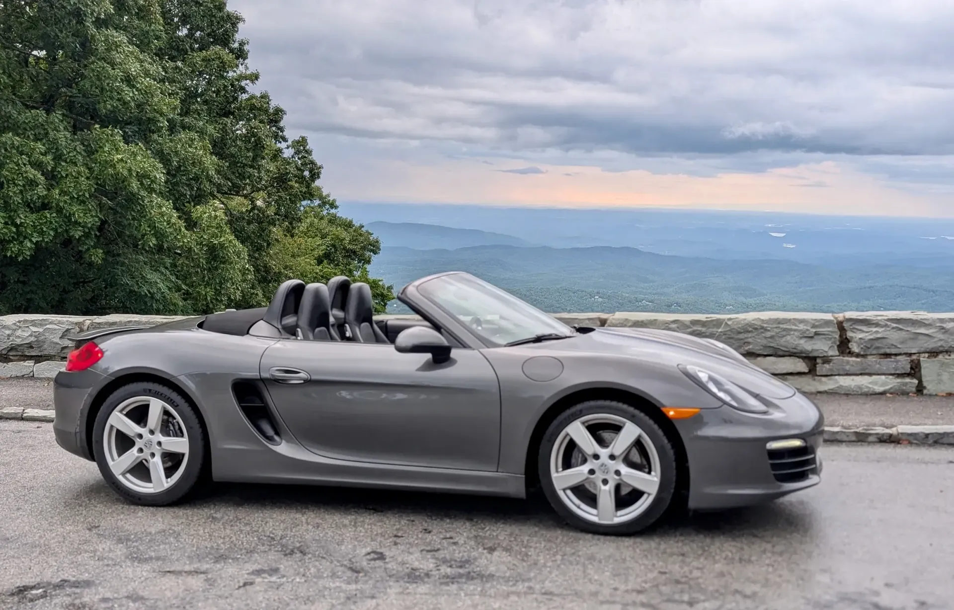 Gray Porsche Boxster convertible parked on a mountain overlook with a cloudy sky and scenic vista.