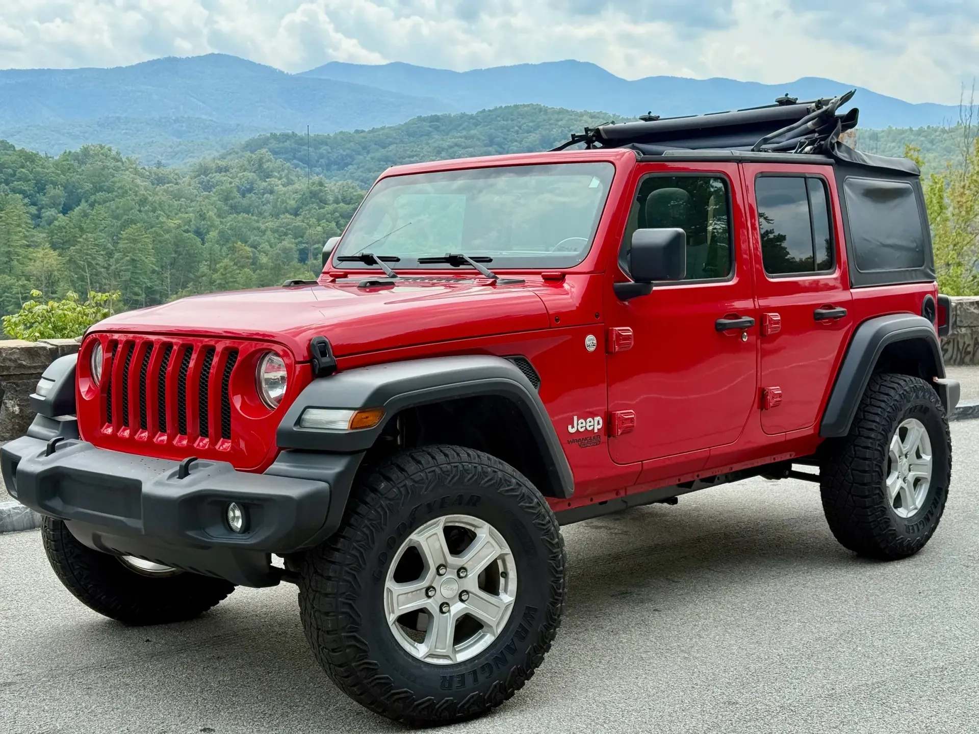 Red Jeep Wrangler parked on a road, mountains in the background, black roof rack.