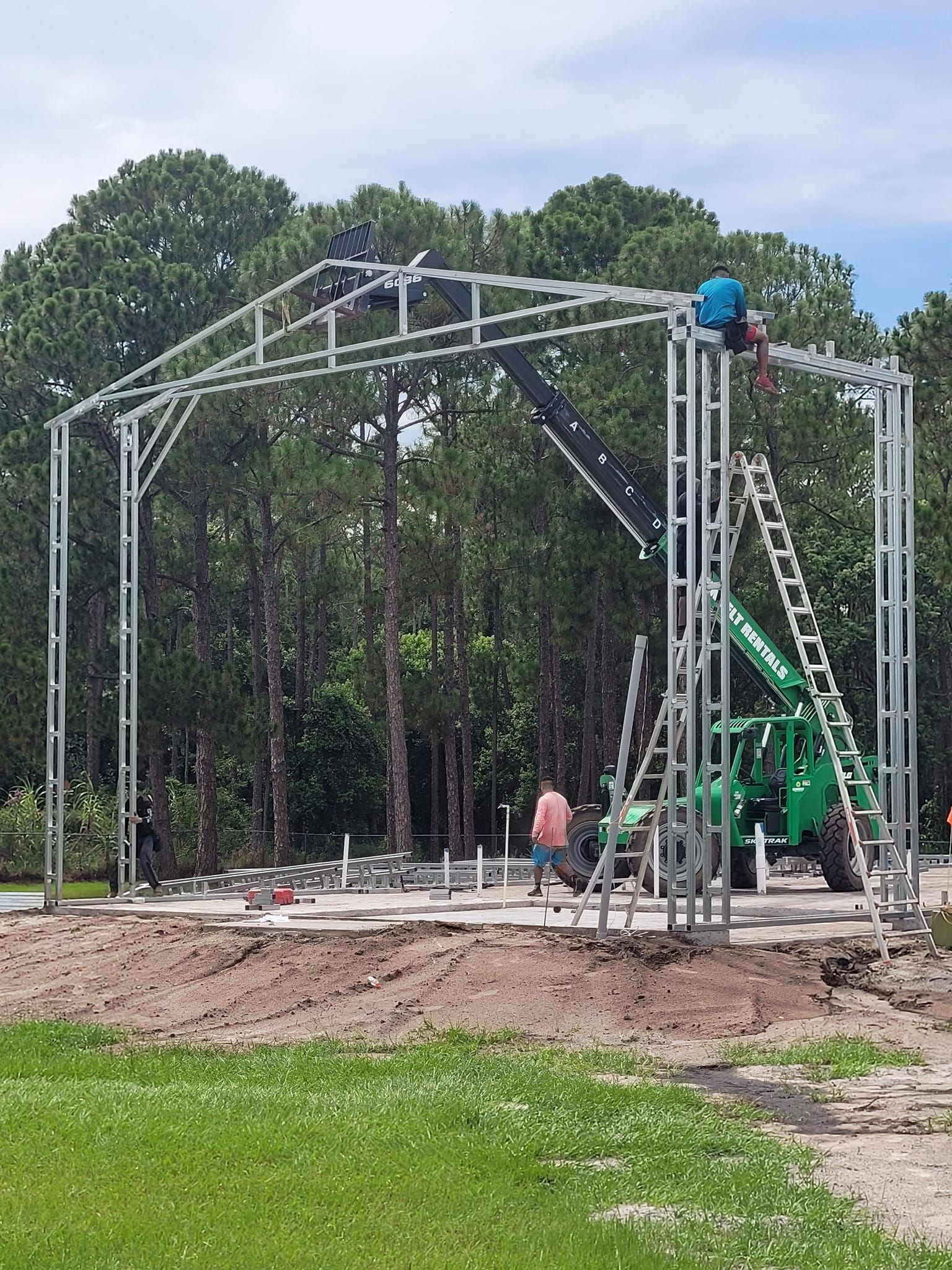 A large metal structure is being built in a field with trees in the background.