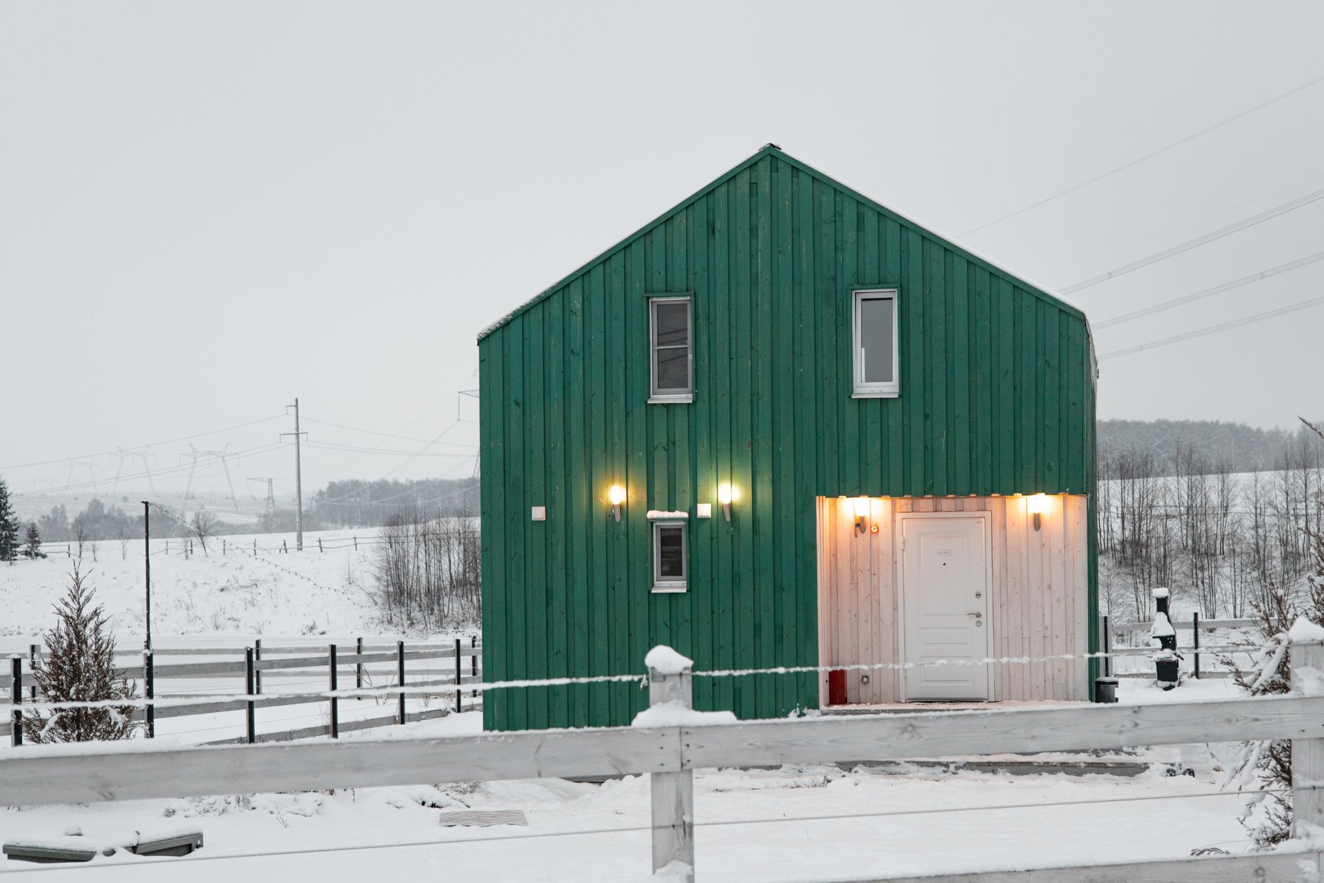 A green barn is surrounded by snow and a white fence